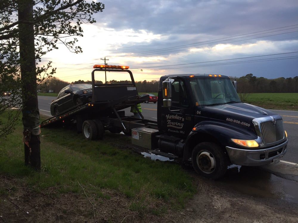 Tow truck loading a dark car by a roadside. Orange flashing lights, cloudy sky, green grass.