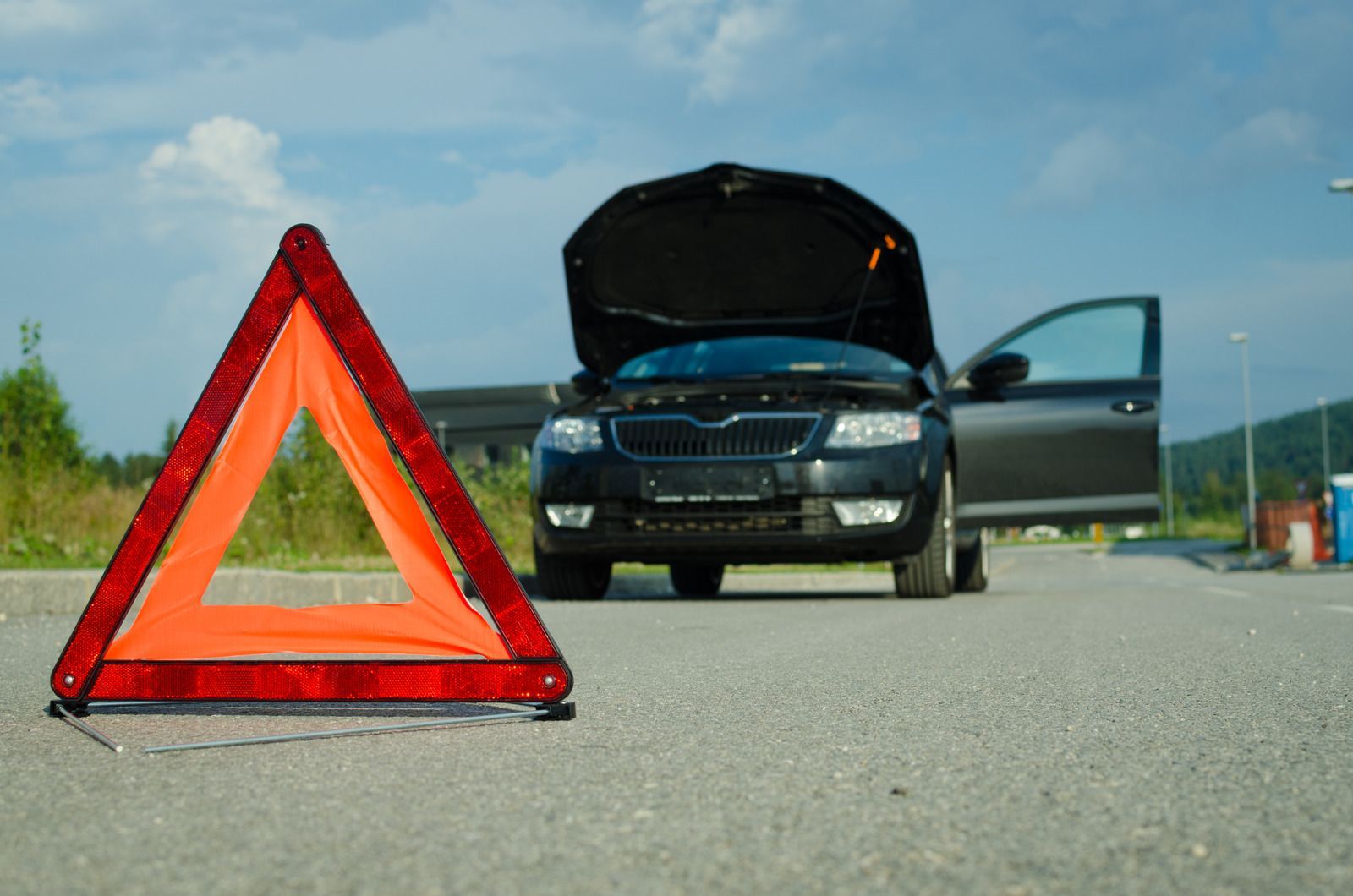 Red triangle warning sign on asphalt in front of a disabled black car with its hood up, outdoors.