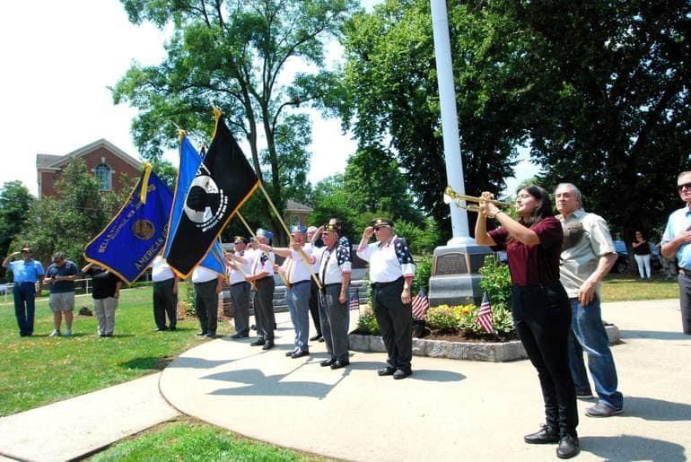 Memorial Day ceremony. People saluting flags. A person plays a bugle near a flag pole and memorial. Green grass and trees.