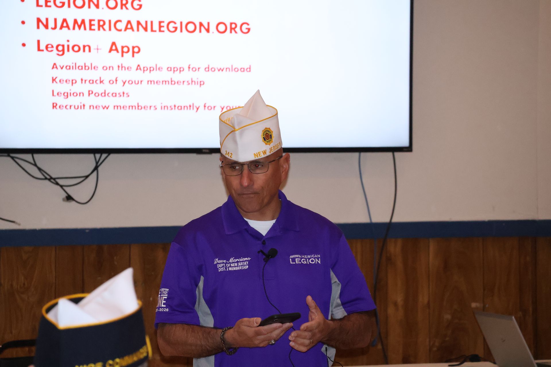 Man in uniform speaking at a podium, holding a phone. A screen displays text.