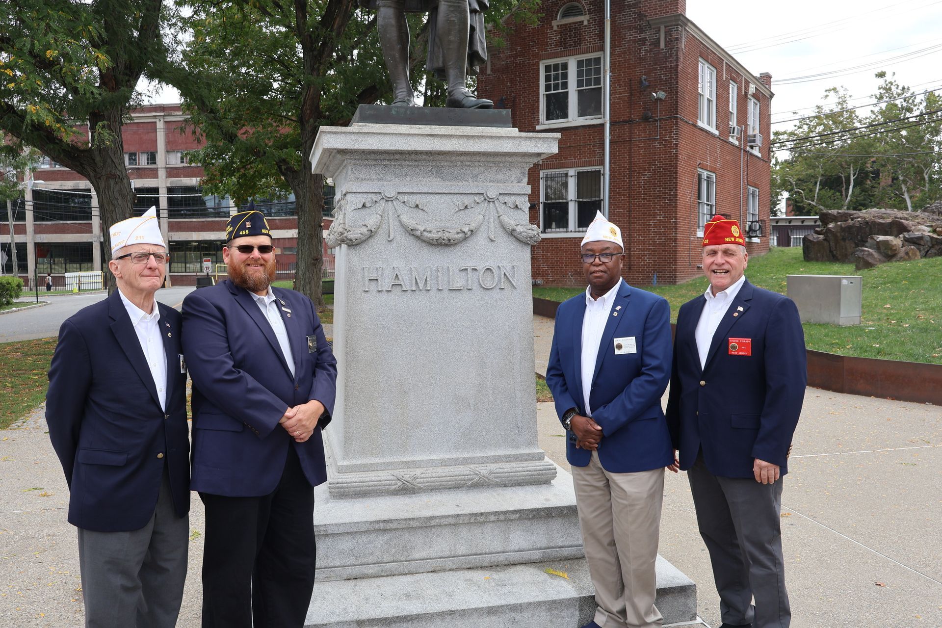 Four men in jackets and white hats stand by a statue of Alexander Hamilton; brick building in the background.