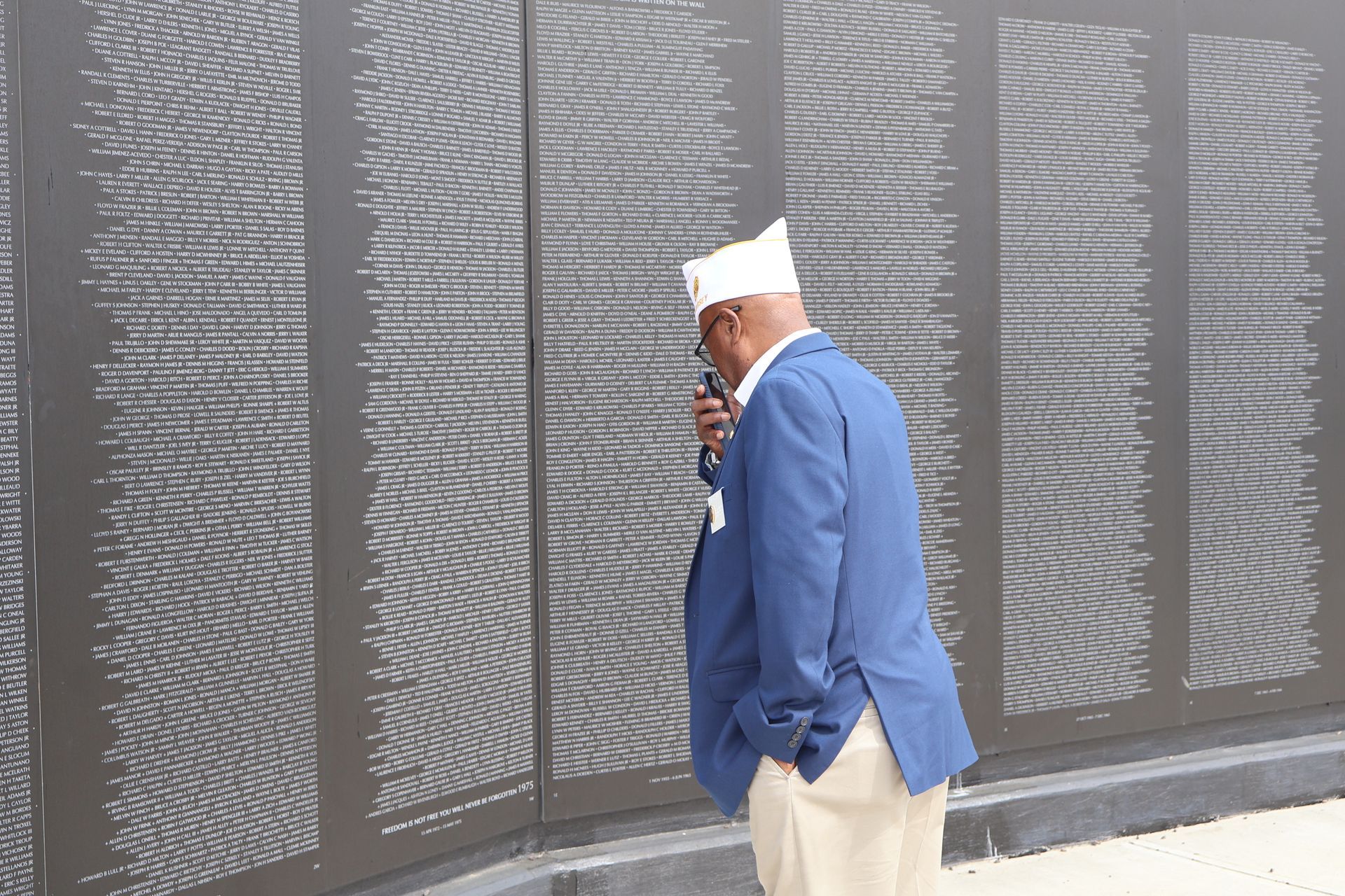 Man in blue jacket looking at a wall etched with names. White hat, microphone visible.