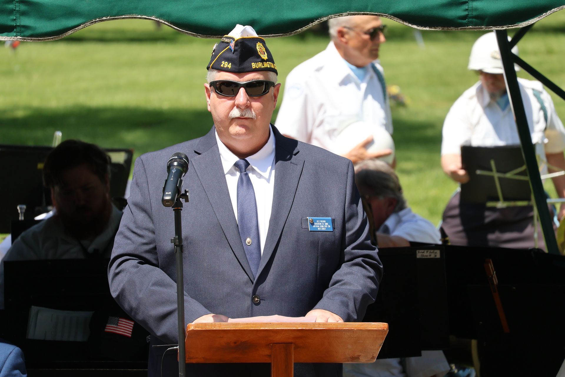Veteran in uniform speaking at a podium outdoors, with microphone and band members in the background