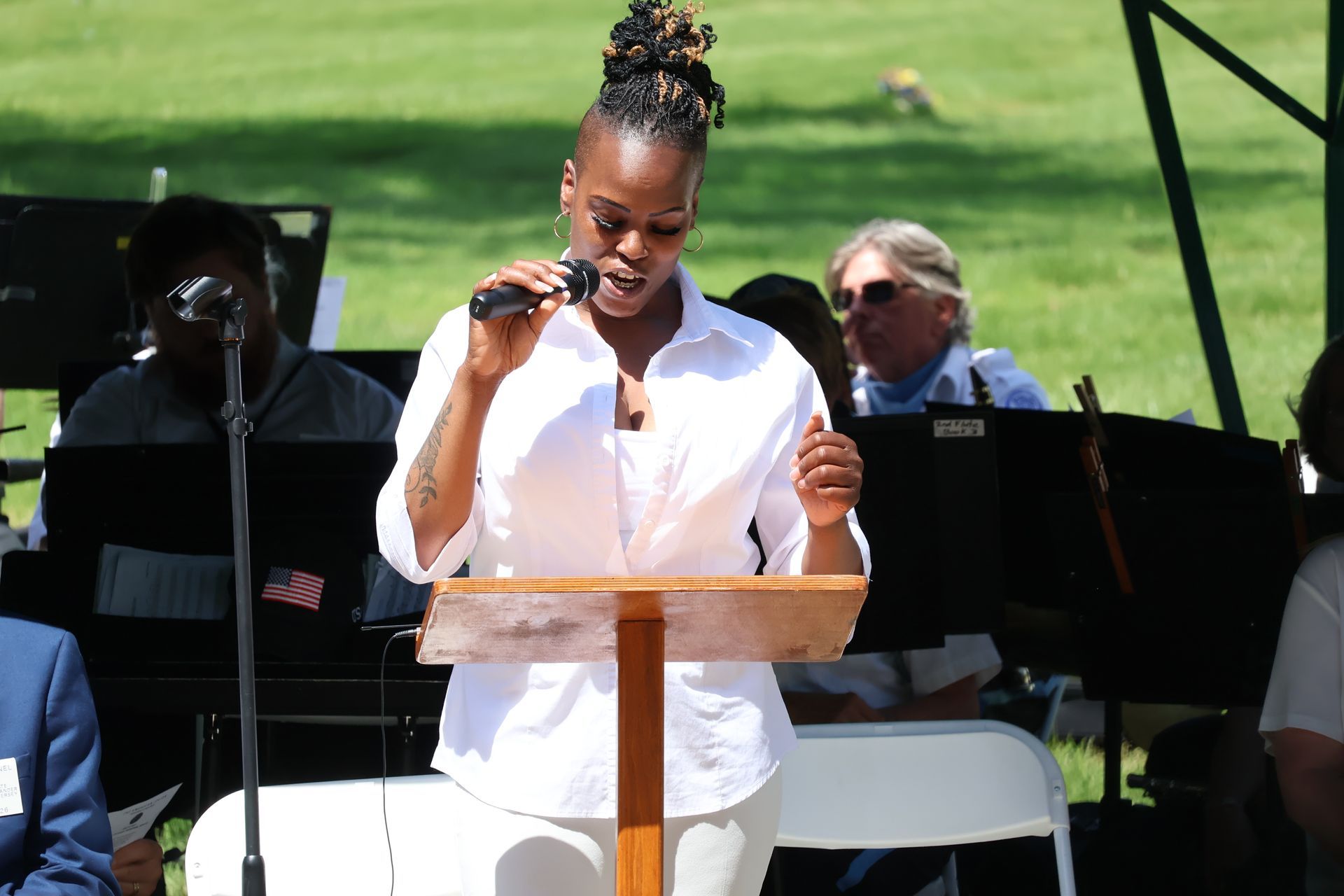 Woman speaking at a podium outdoors, with seated musicians in the background
