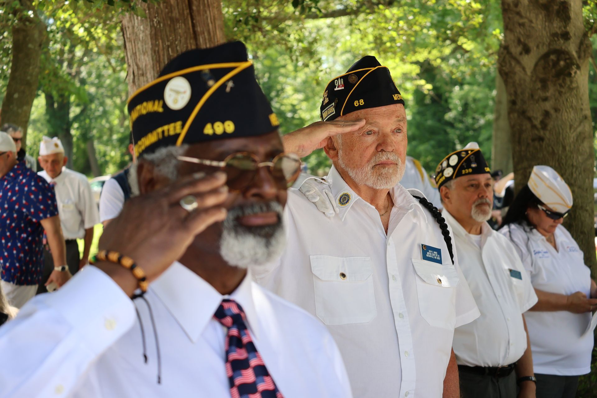 Veterans in white uniforms and black caps stand outdoors under trees during a ceremony