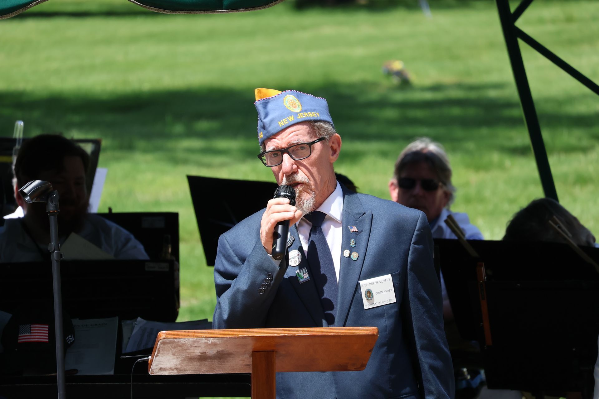 Man in blue cap speaks at podium outdoors, holding a microphone during a ceremony