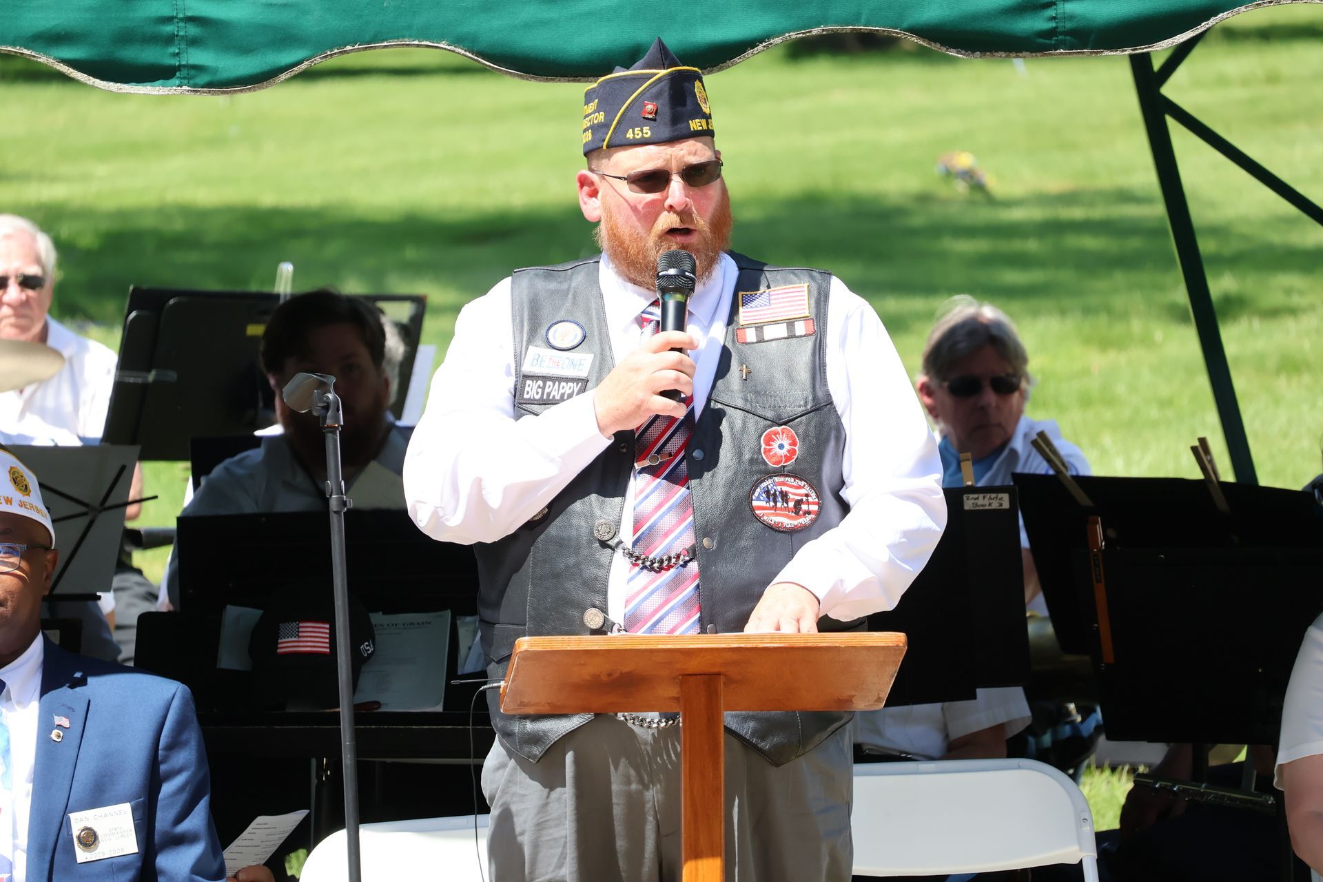 Man speaking at a podium outdoors, wearing a vest and hat, with musicians seated behind him.