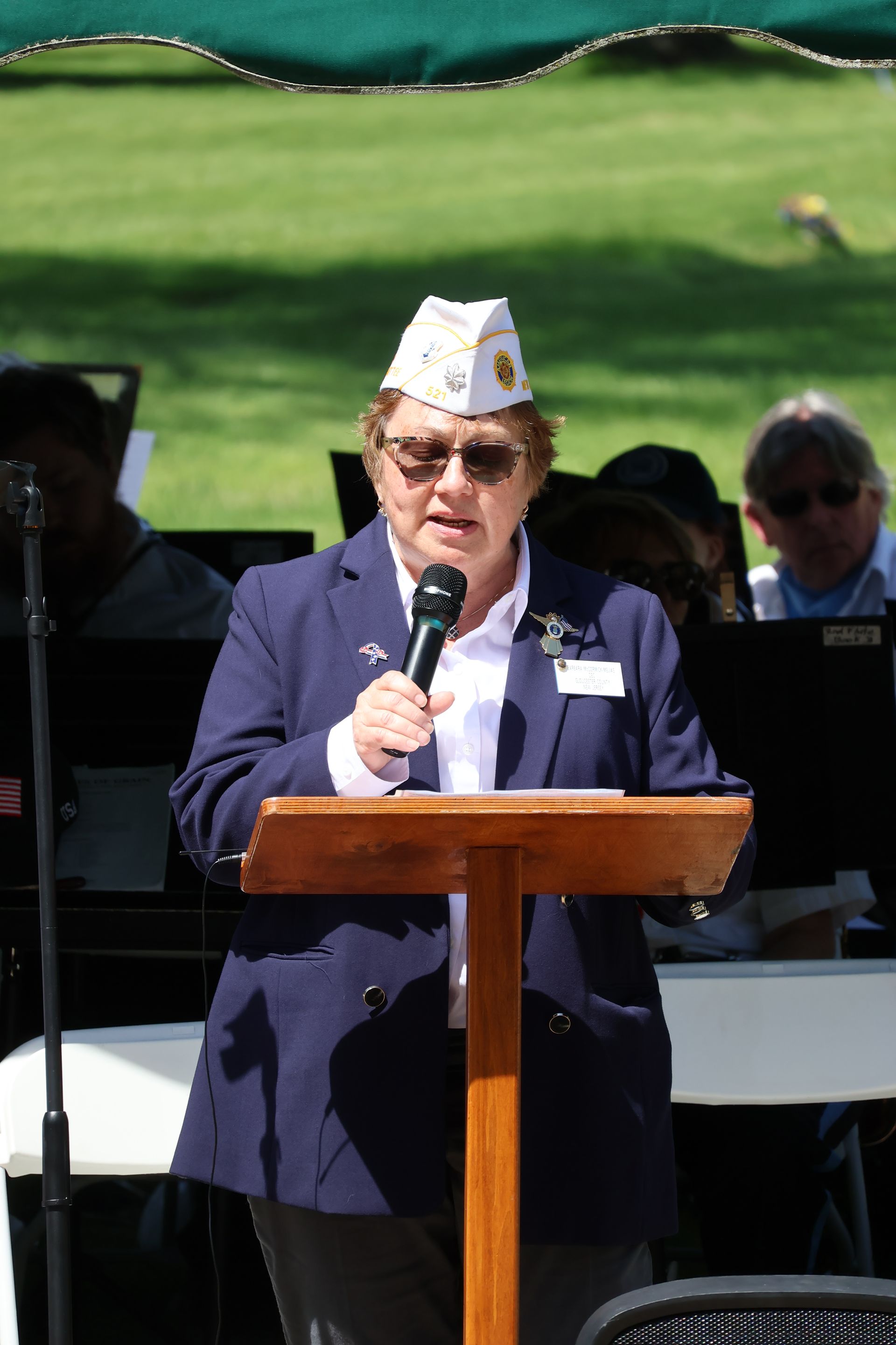 Person speaking at a podium outdoors, wearing a navy suit and paper hat, with musicians seated behind.