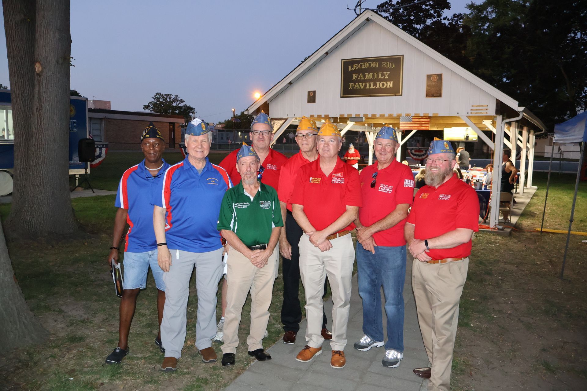 Group of men in caps and red shirts pose in front of a small building.