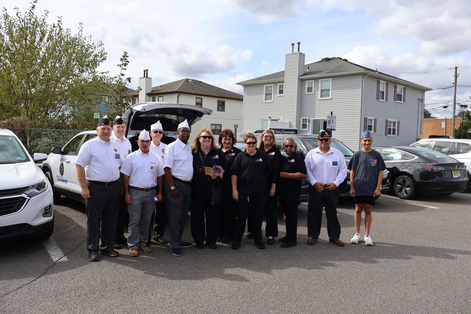 Group of people in uniform standing in front of cars, likely at a memorial or event, outdoors.