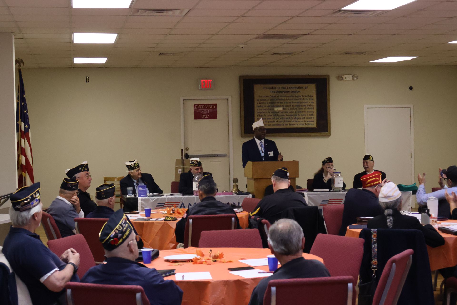 Man speaking at a veterans' meeting, many in attendance. US flag, orange tablecloths, neutral-toned walls.