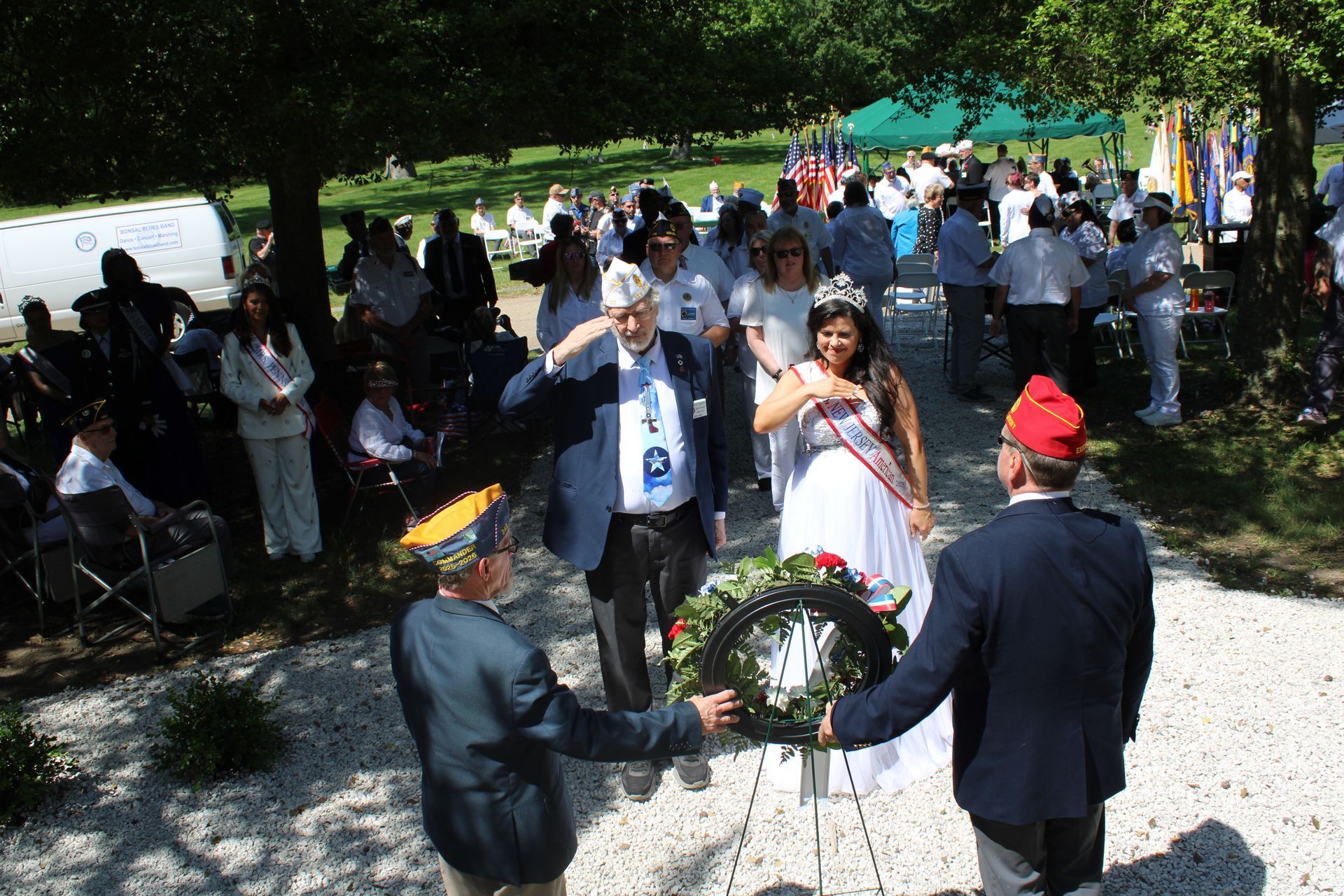 People gathered outdoors at a wreath-laying ceremony, with a red-capped man near a memorial wreath.
