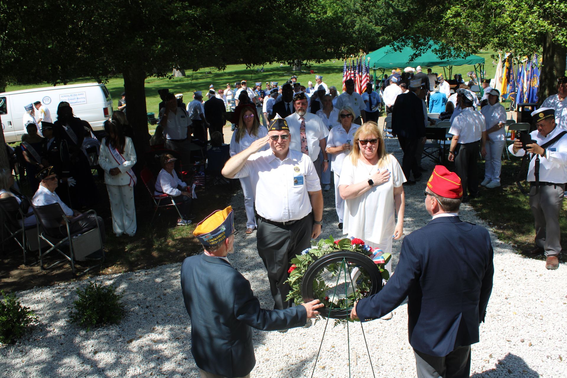 People gathered outdoors around a wreath ceremony, with two men holding the wreath in the foreground.