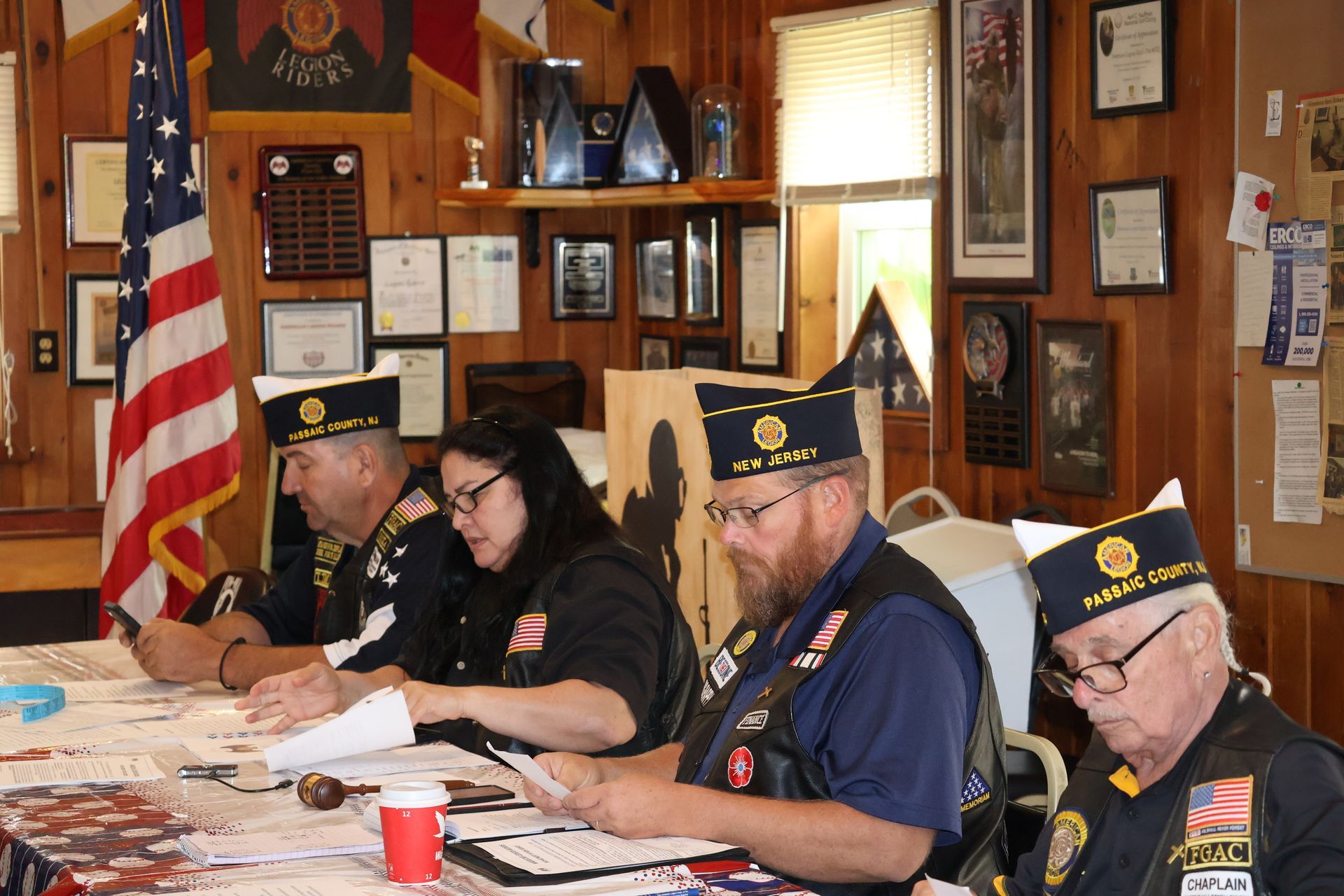Four people wearing veteran attire sit at a table reviewing documents, American flag in background.