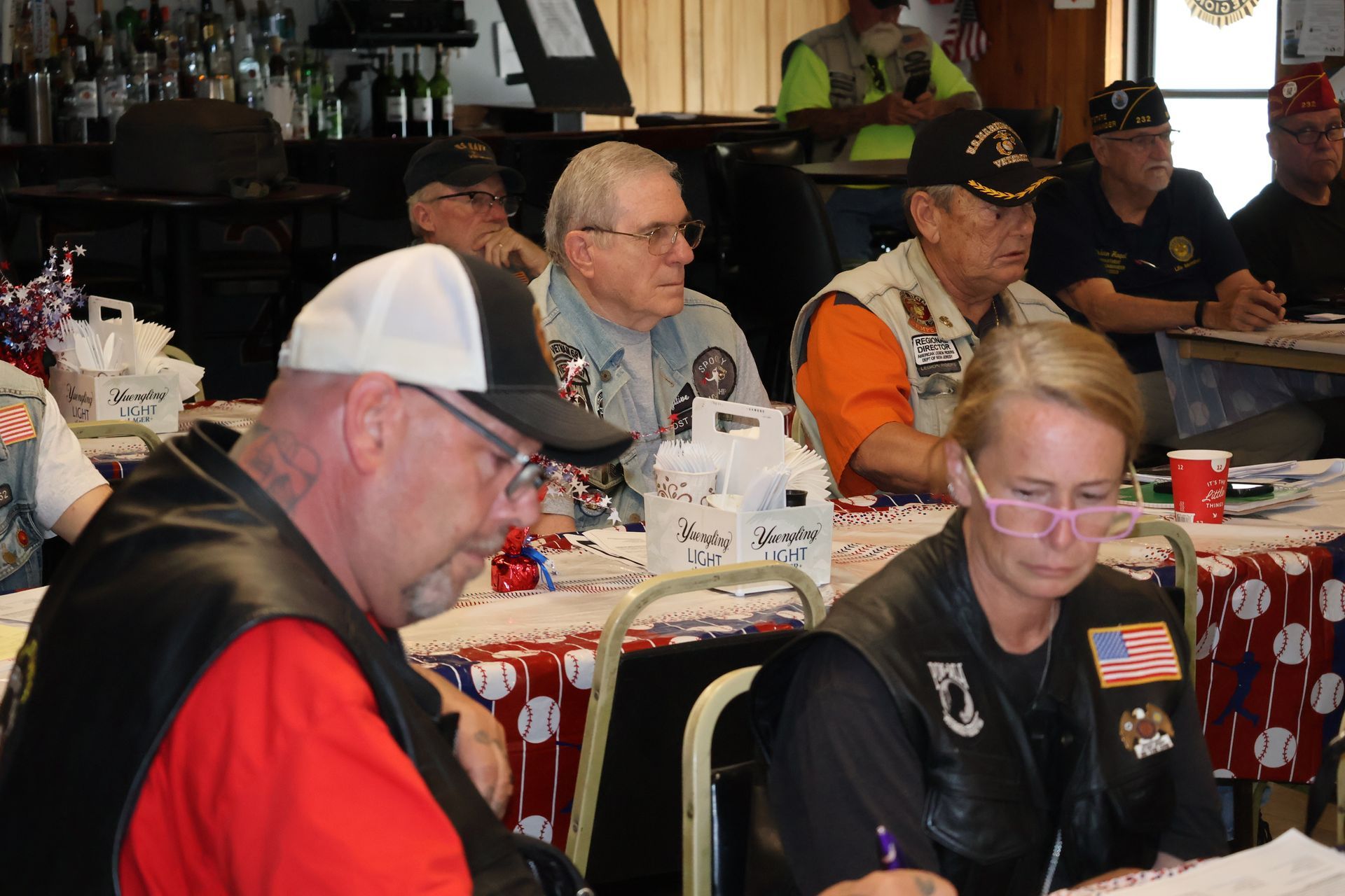Veterans seated at tables in a room, some wearing vests and hats, possibly at a meeting or event.
