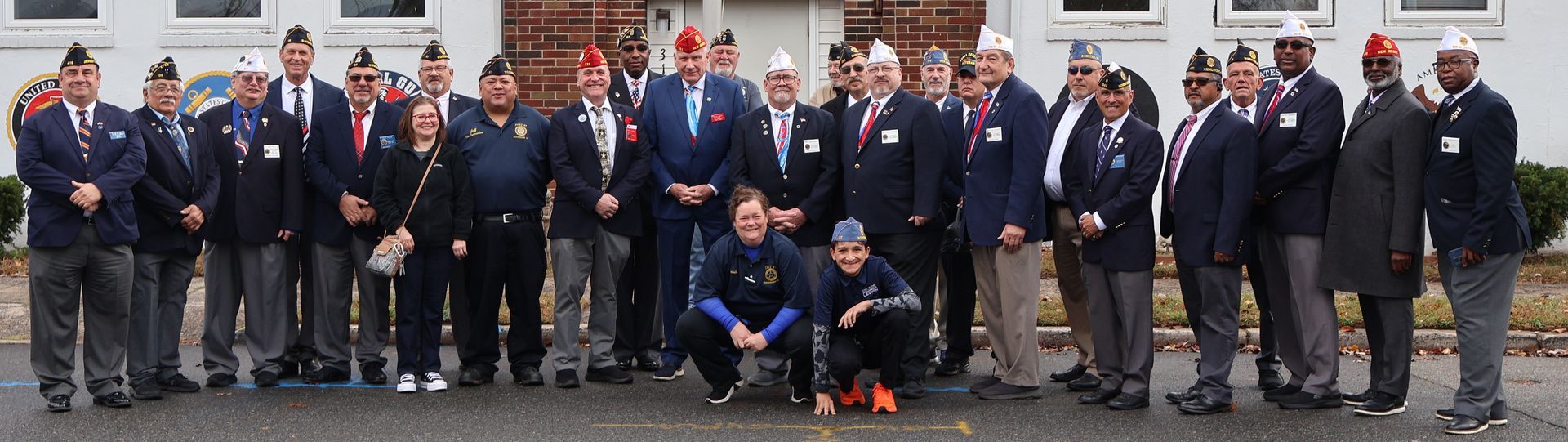 Group of people in matching blue blazers, some wearing caps, standing in front of a building.