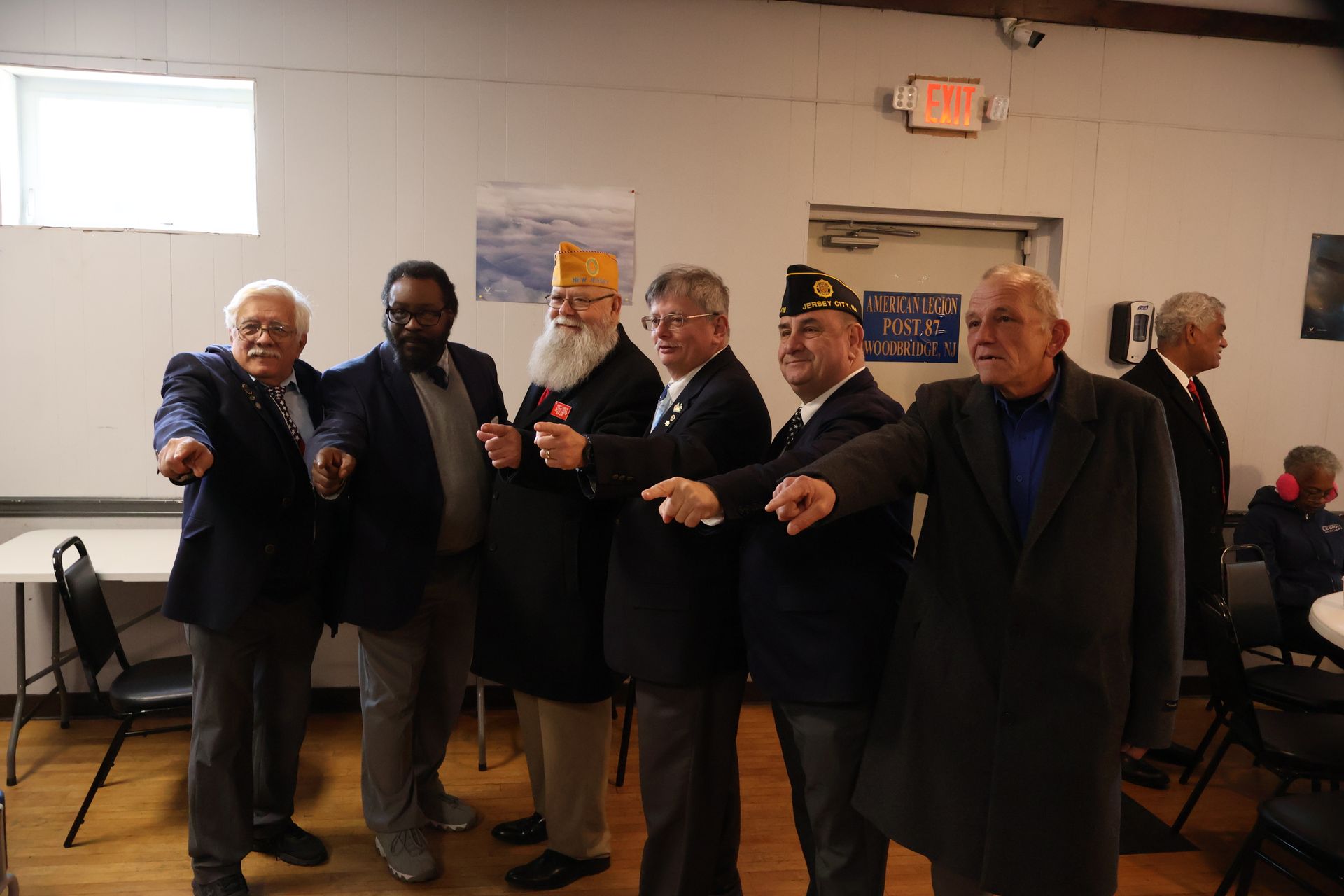 A group of men in suits pointing. Indoors with tables, an exit sign and a small painting are visible.