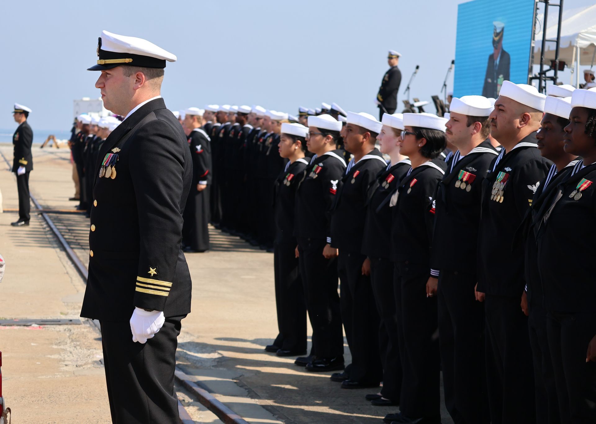 Naval officers and sailors in dark uniforms stand at attention on a pier with water in the background.