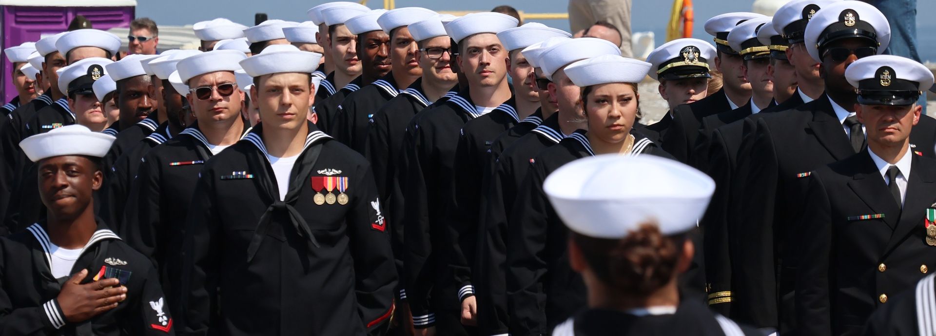 Sailors in navy uniforms and white hats stand outdoors, some with hands over their hearts.