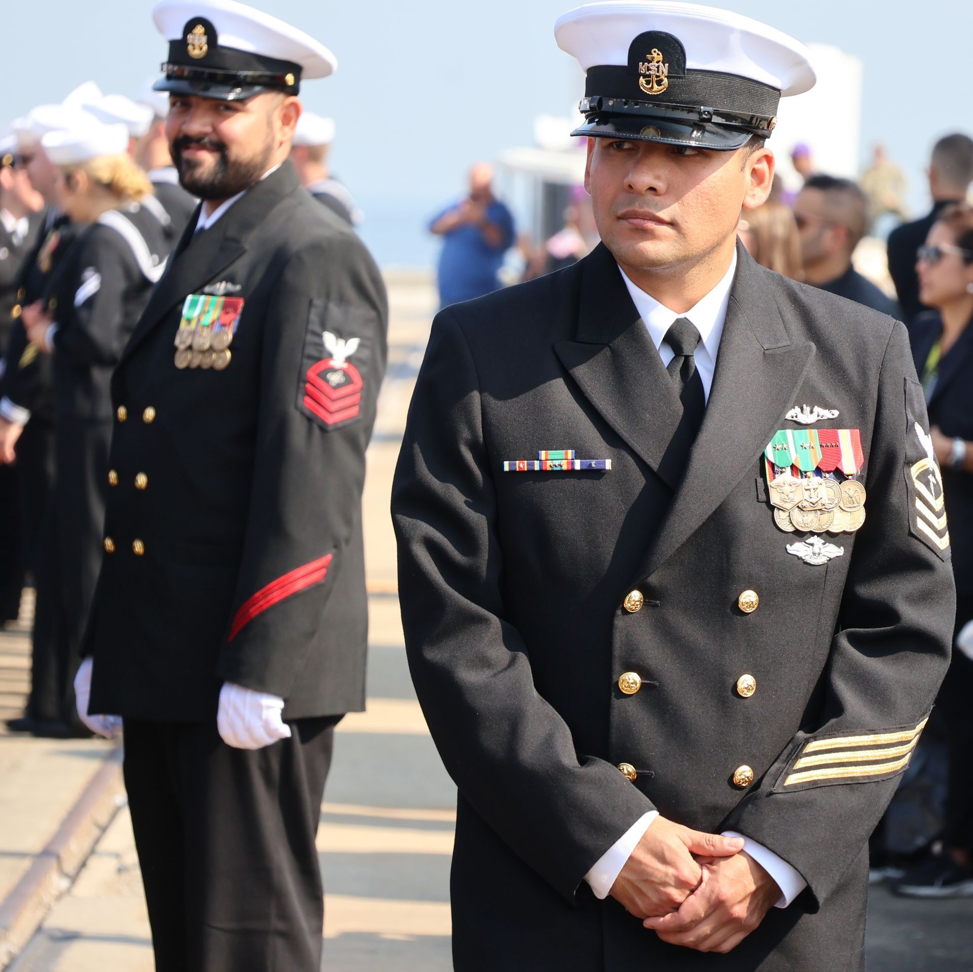 Two U.S. Navy officers in dress uniforms standing outdoors; one with folded hands, other looking to the side.