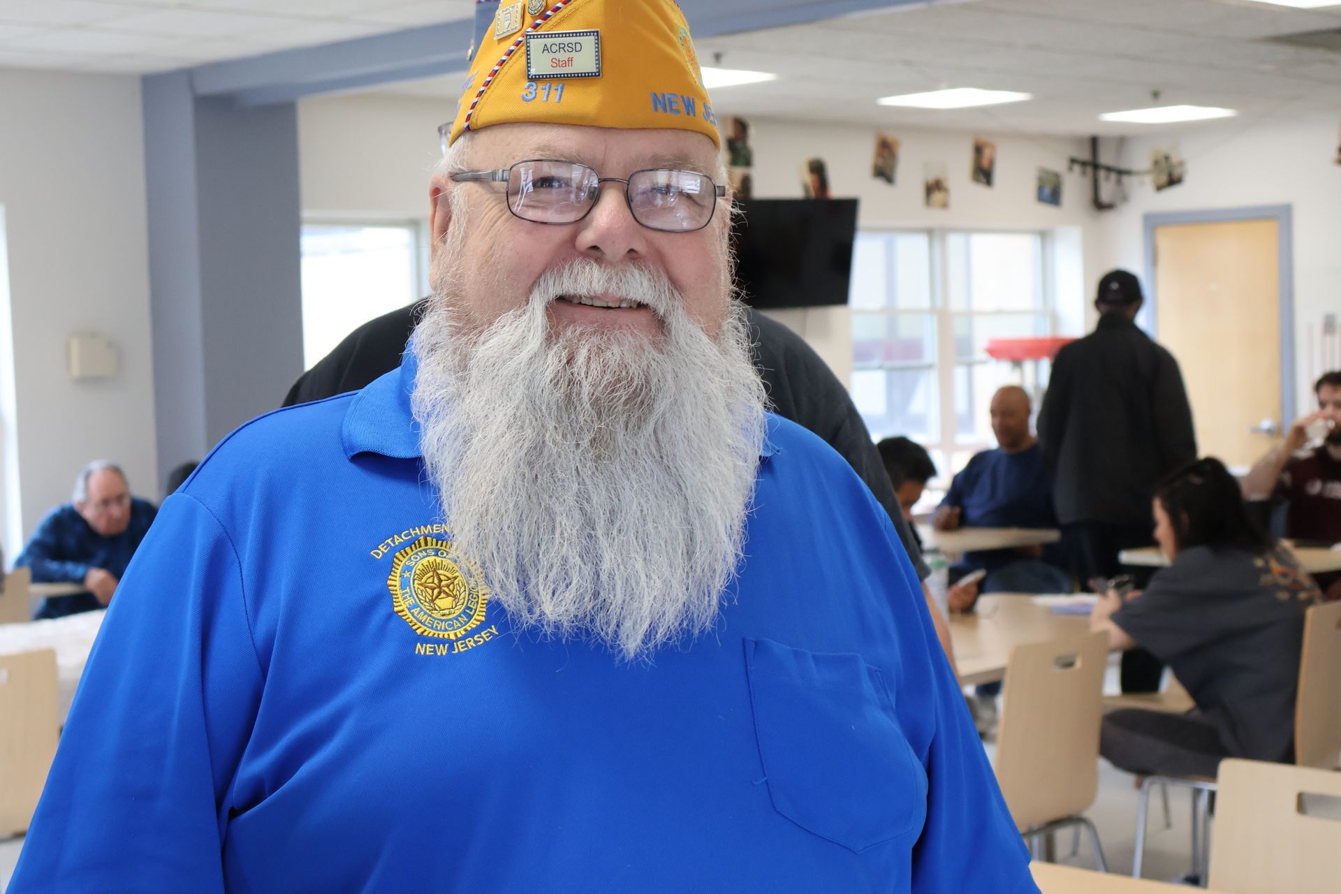 Man in blue shirt and veteran cap smiles, indoor cafeteria setting.