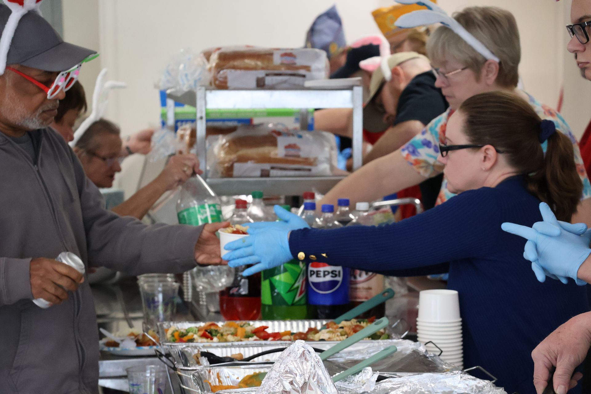 People at a food service line, one is served a cup. Beverages and dishes are on the table.