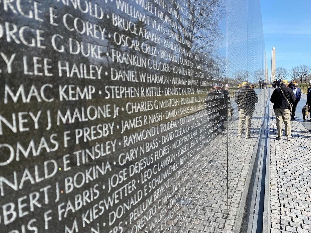 Vietnam Veterans Memorial with engraved names. People are viewing the wall. Washington Monument in background.