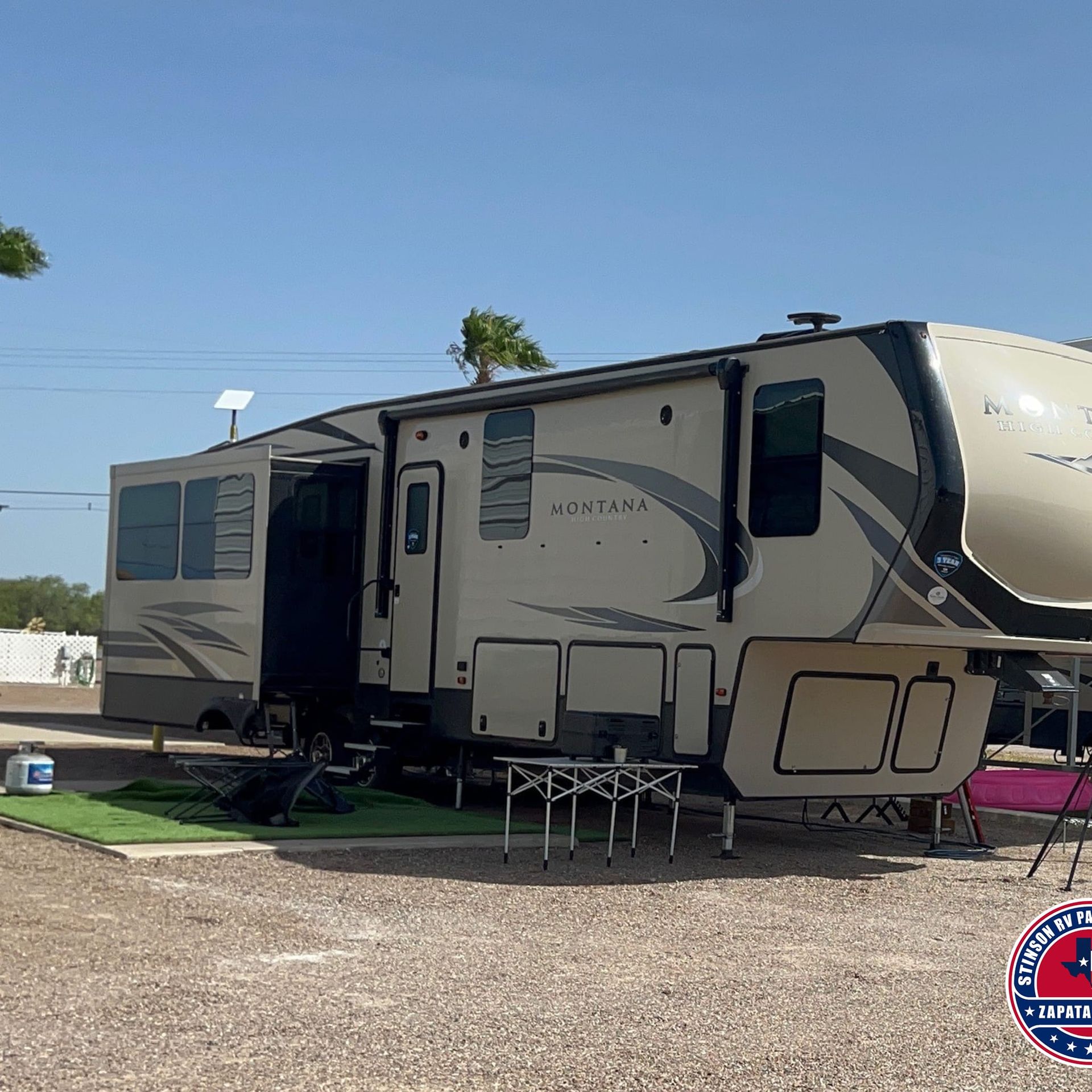 Tan RV with slide-out extended in a gravel campsite. Blue sky, palm trees.