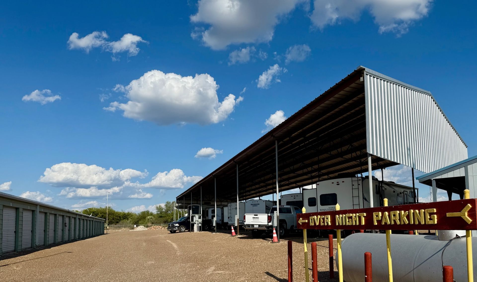 RV parking under a metal roof, with storage units, gravel ground, and a blue sky.