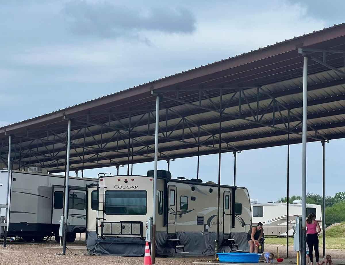 RVs parked under a metal awning on a cloudy day. Two people sit outside one RV.