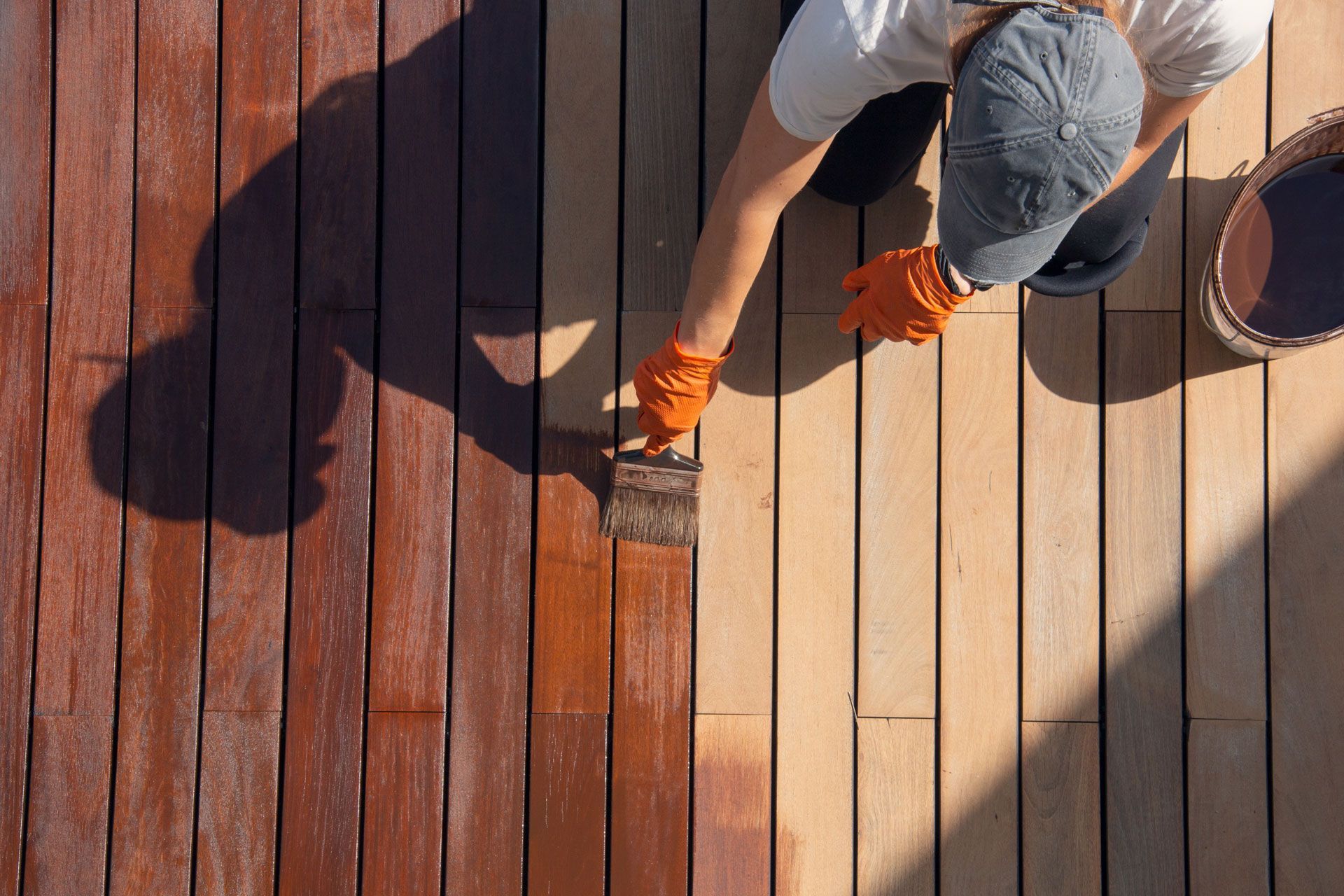 Person staining wooden deck with brown stain; a bucket and shadow are visible.