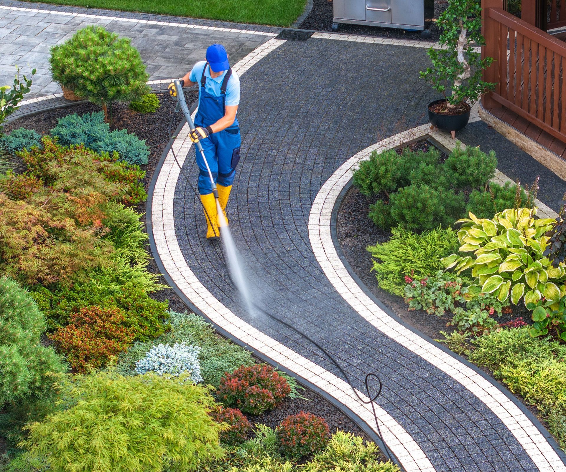 Person power washing a black cobblestone pathway with white trim, surrounded by greenery.