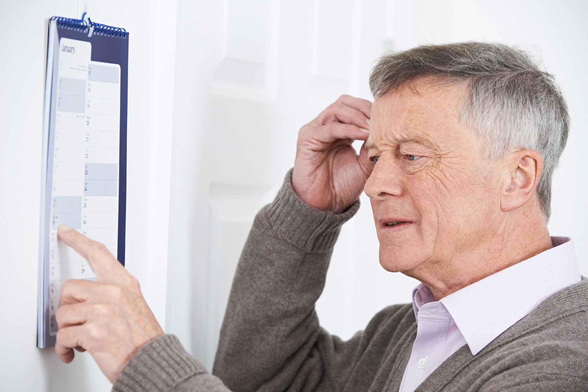 Elderly man looks at wall calendar, hand on head.