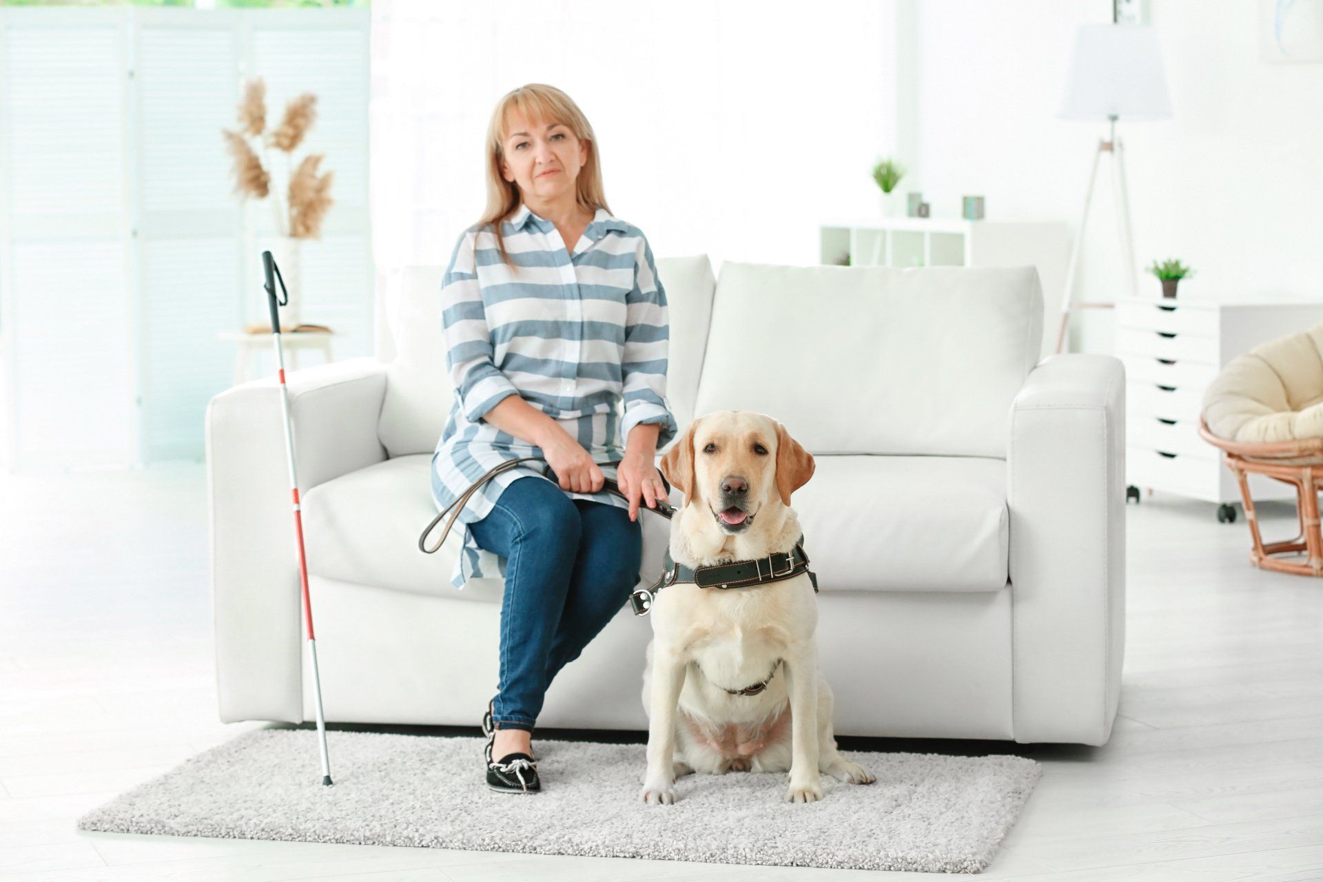 Woman with guide dog sitting on a couch, a cane beside her in a bright living room.