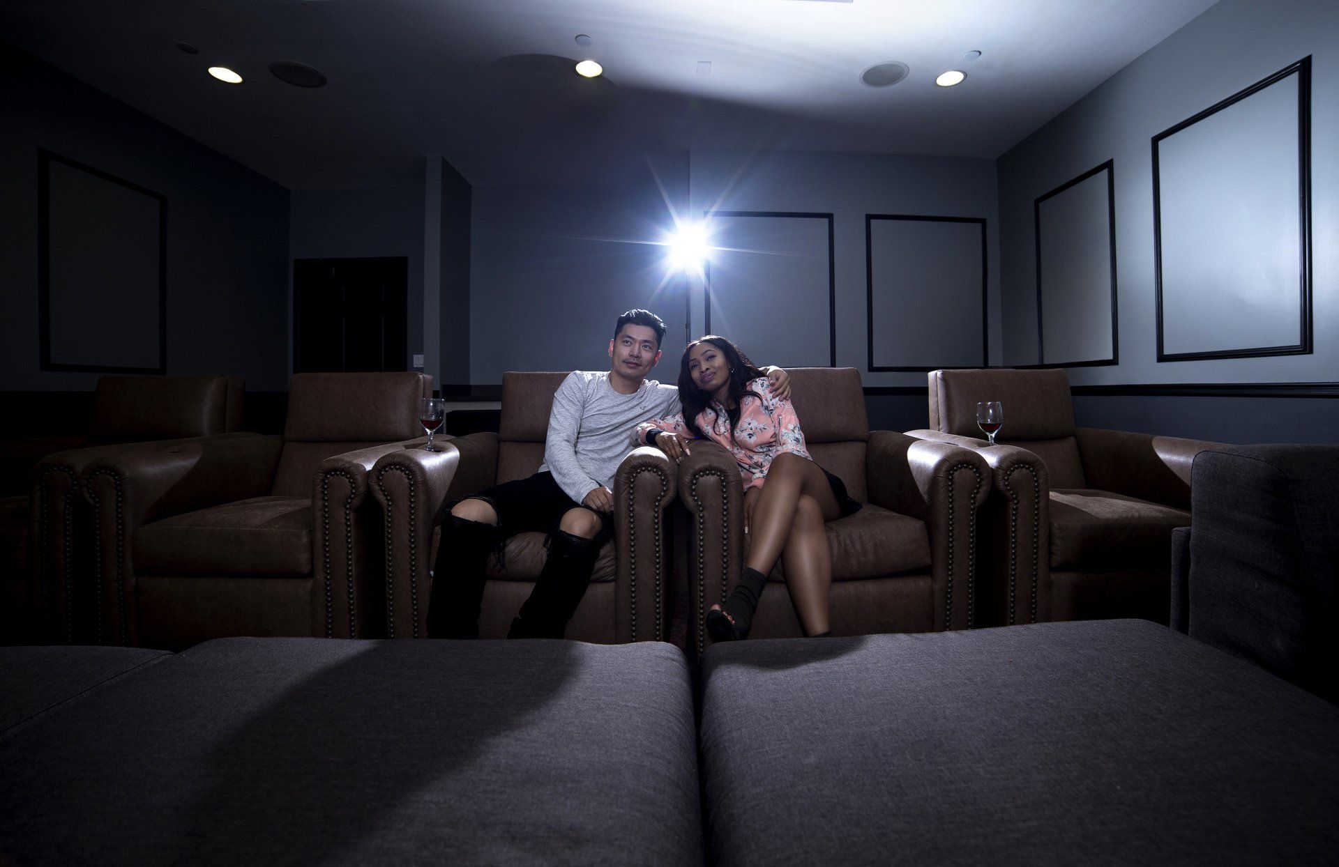 A photograph of a male and female young couple, sat on some leather sofas in a private home cinema, watching a movie