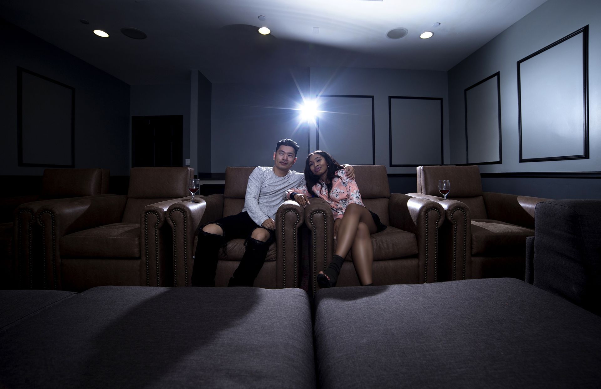Two people in a home theater, seated on a couch. A bright light shines, and empty screens are on the wall.
