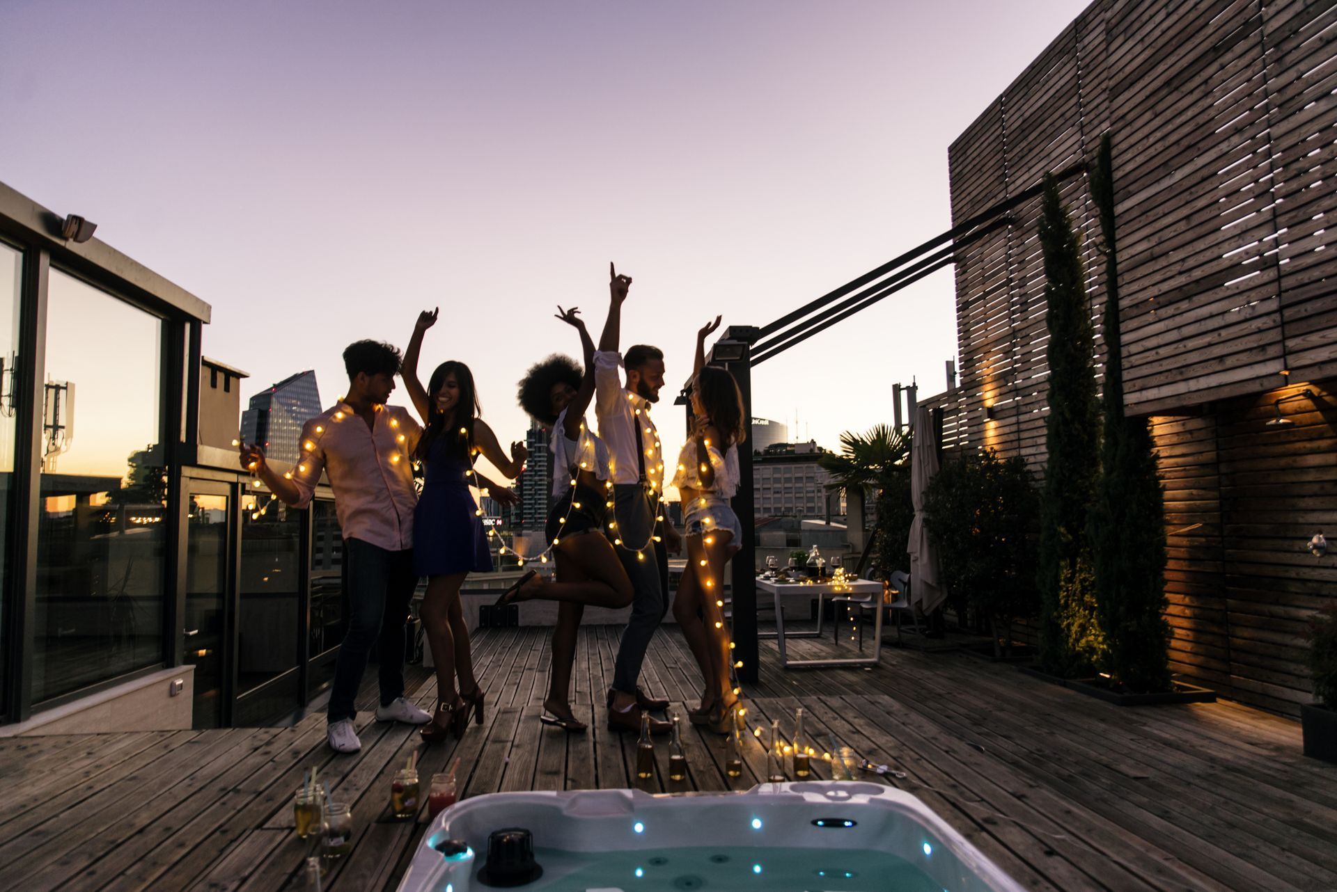 Group dancing on a rooftop patio at dusk. Fairy lights, hot tub, city skyline.