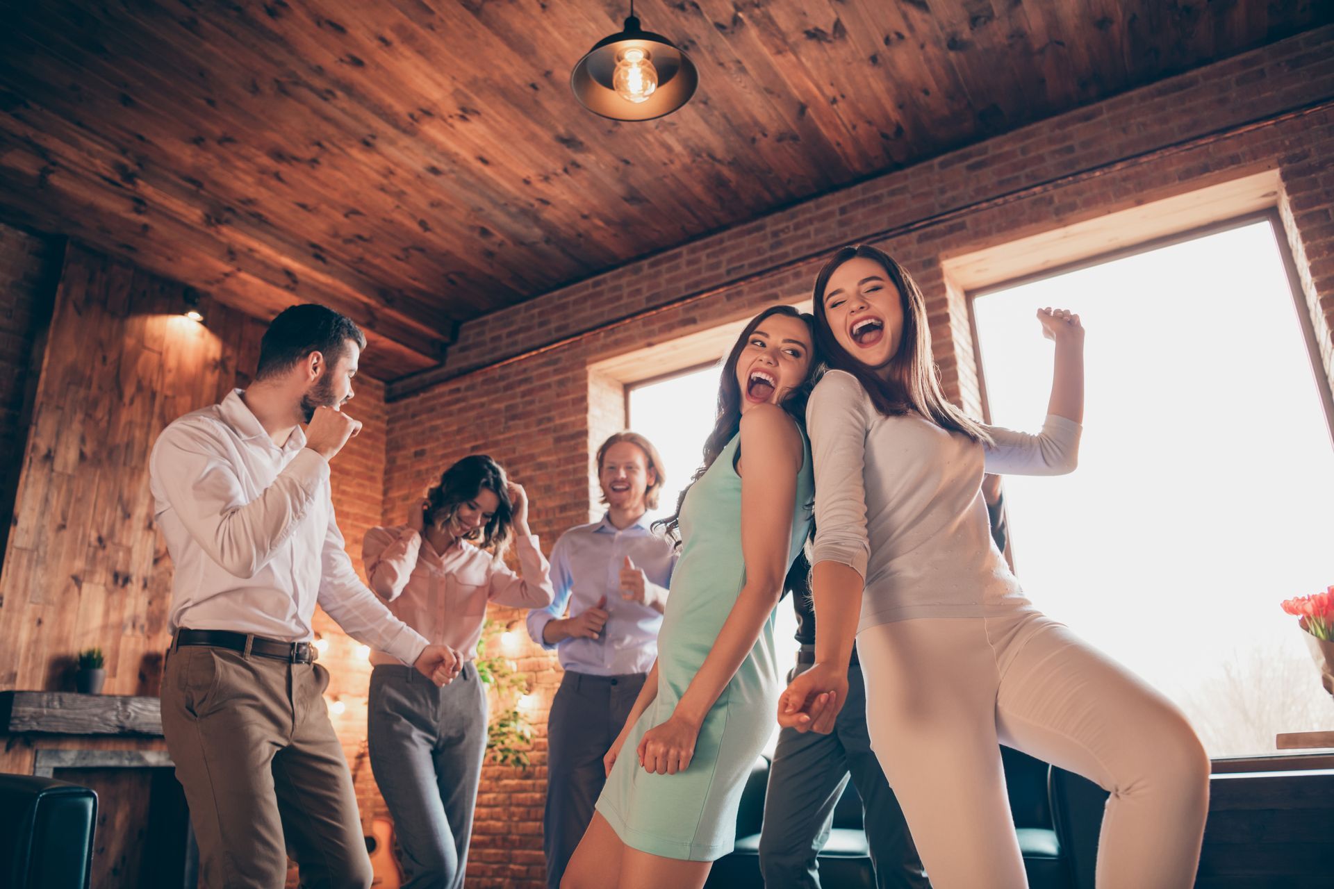 People dancing joyfully in a room with wooden walls, natural light, and a hanging light.