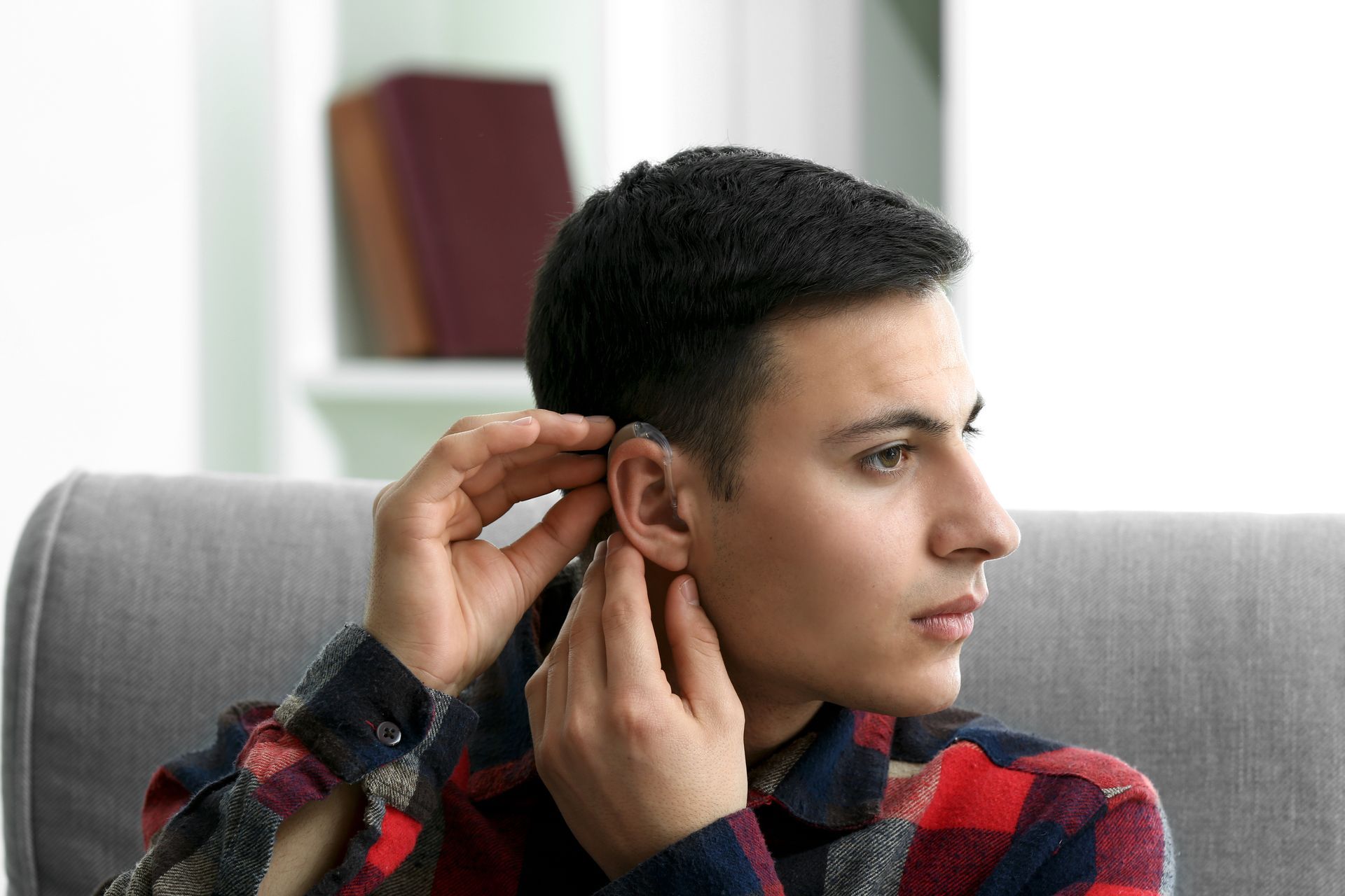 Man adjusting hearing aid, sitting on a couch. He is wearing a plaid shirt, in a room with bookshelves.