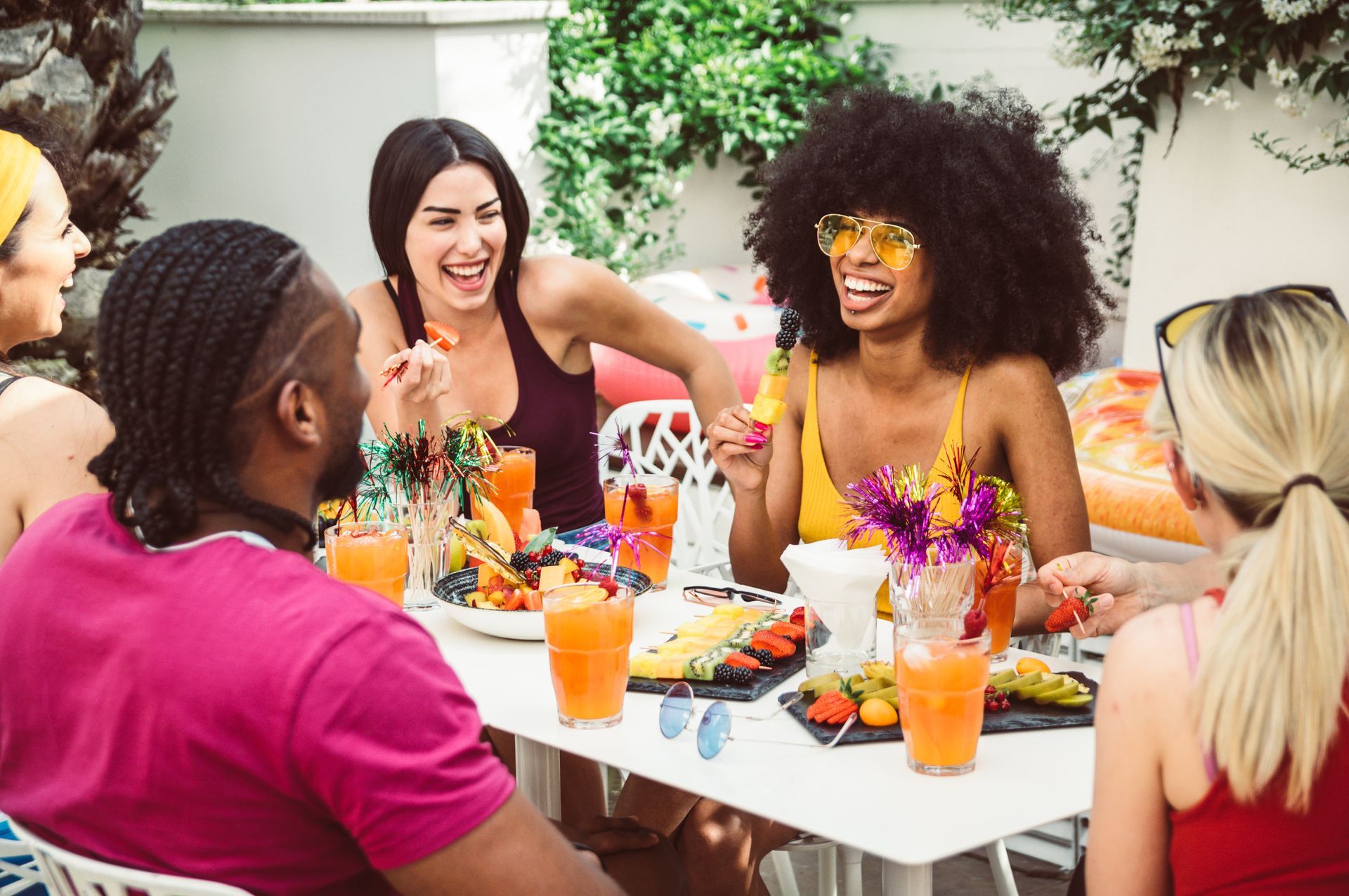 Friends laughing and eating together at an outdoor party, colorful food and drinks on a white table.