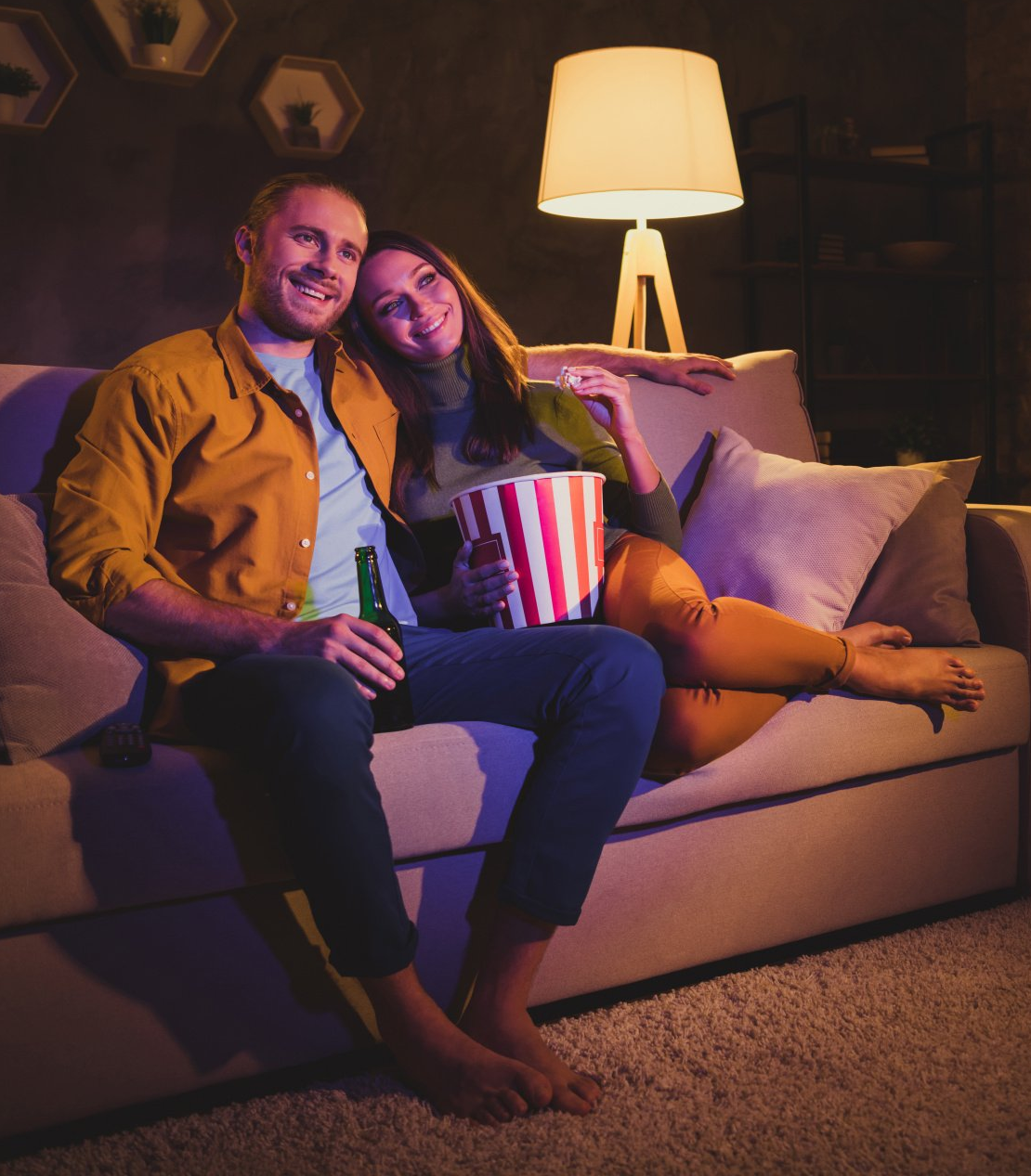A photograph of a male and female couple, sat on a sofa arm in arm, watching a movie while eating popcorn