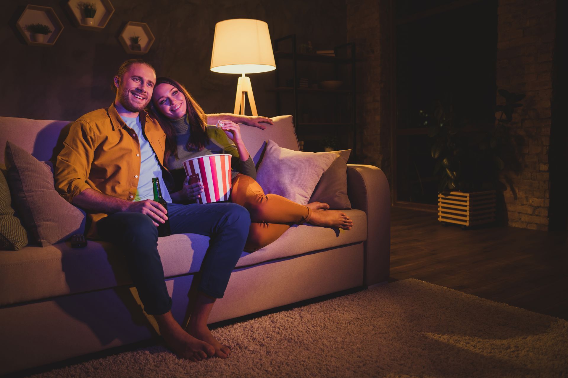 Couple on a couch watching TV, holding popcorn and a drink. Warmly lit, relaxing at home.