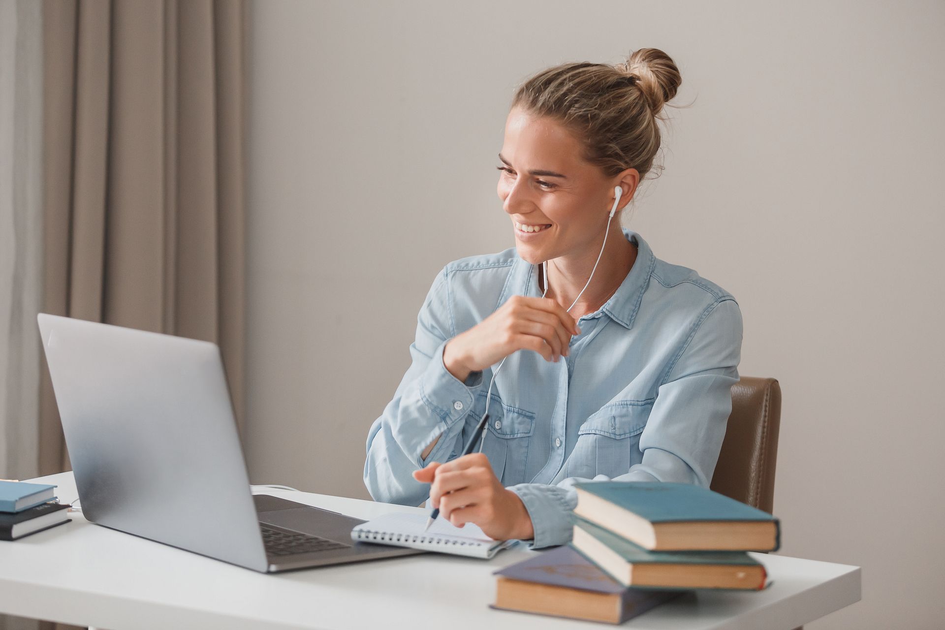 Woman in a light blue shirt, smiling during a video call on a laptop, sitting at a desk.
