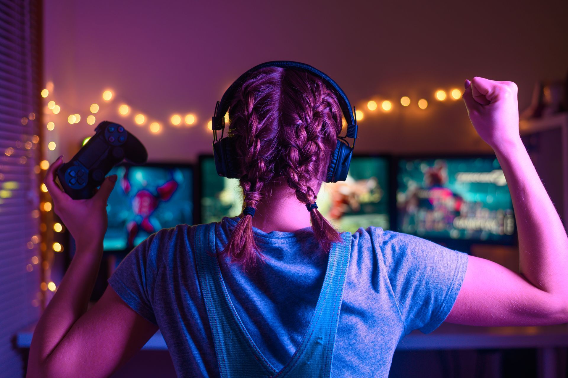 A person with headphones, braids, and a controller celebrates in front of three lit screens, fairy lights visible.