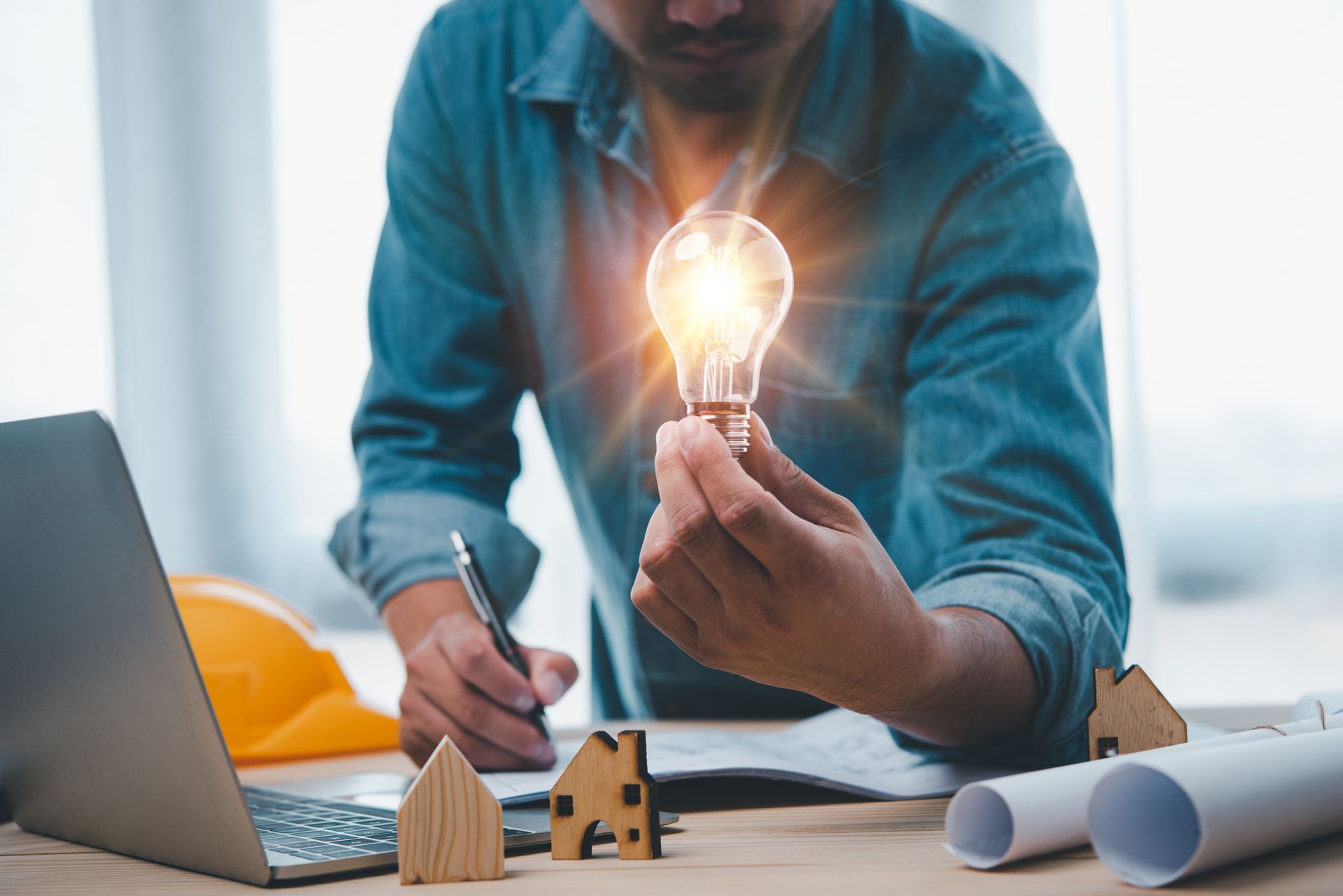Man holding glowing lightbulb, working on laptop and blueprints, surrounded by miniature houses and a hardhat.