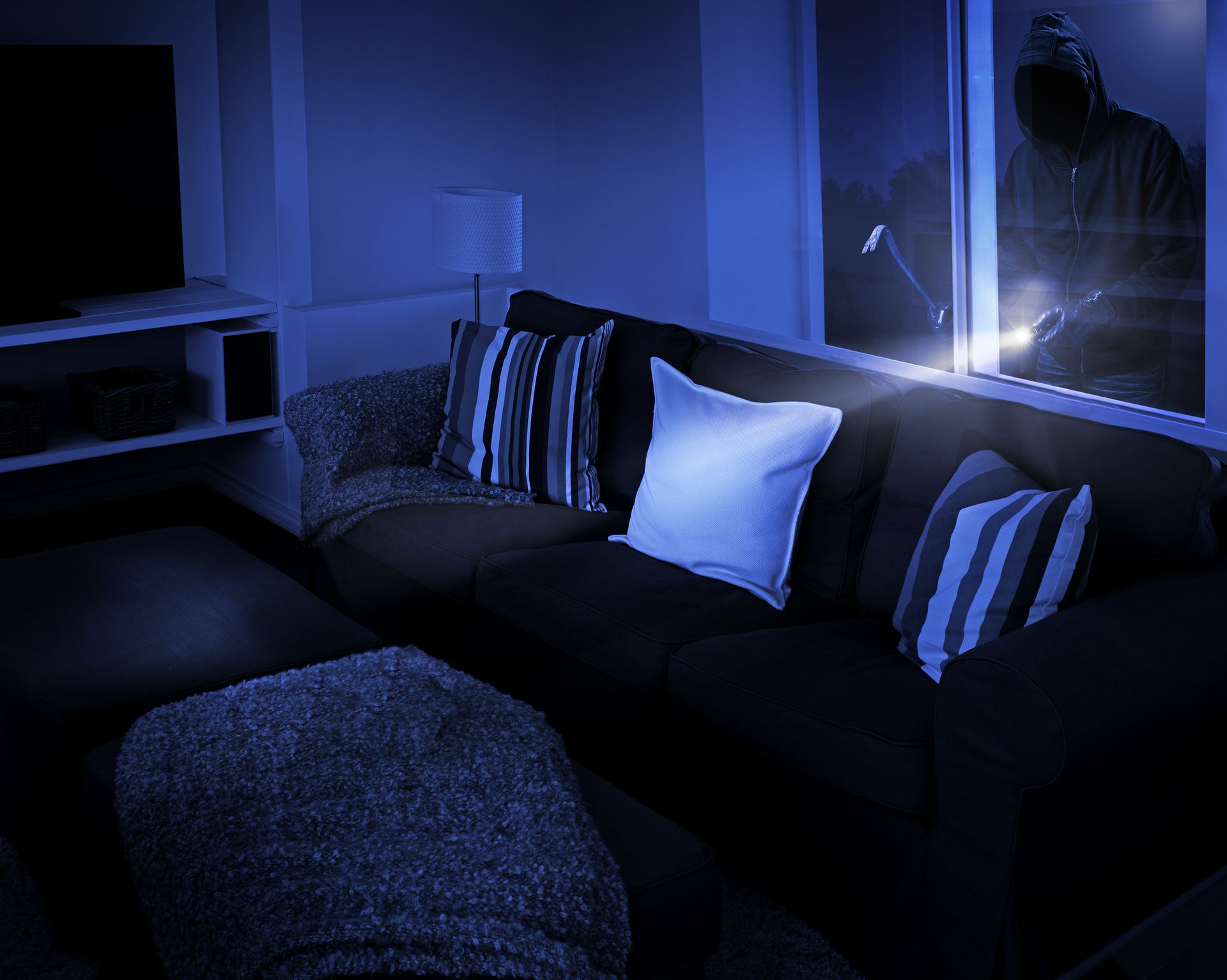 A dark living room. A person in a hoodie breaks into the house through a window at night.