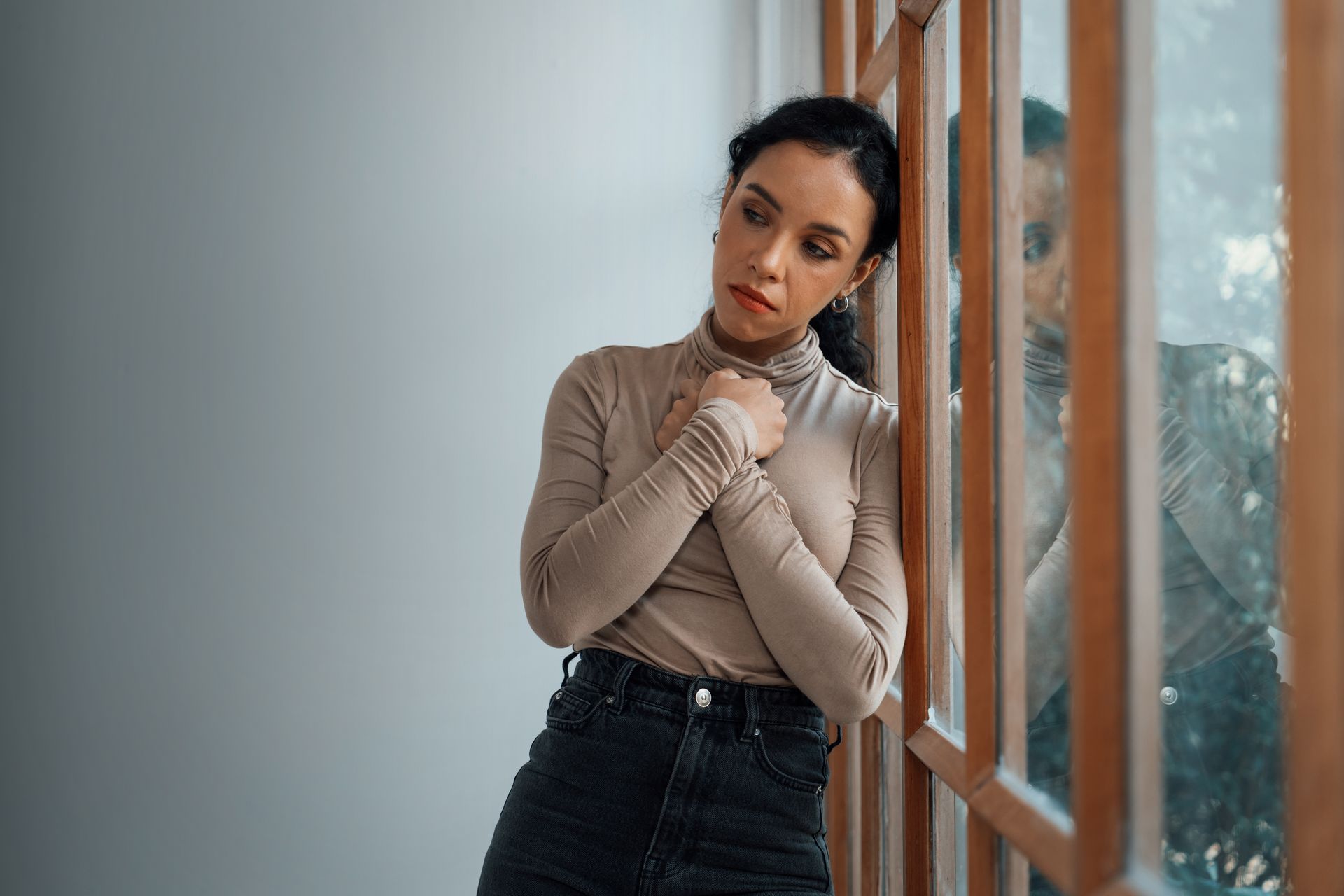Woman in tan turtleneck leans against a window, looking thoughtful with hands clasped.