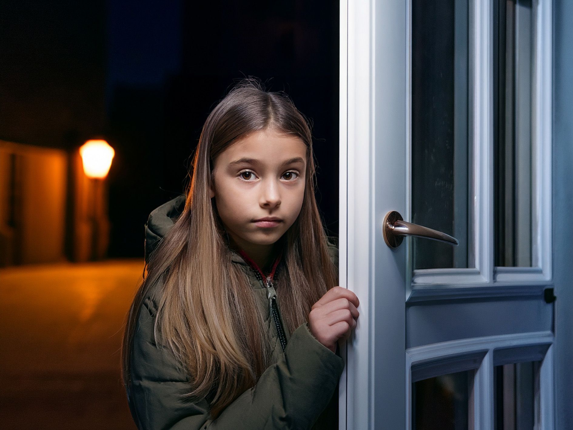Girl at a doorway at night, wearing a green jacket, looking concerned; street lamp in background.