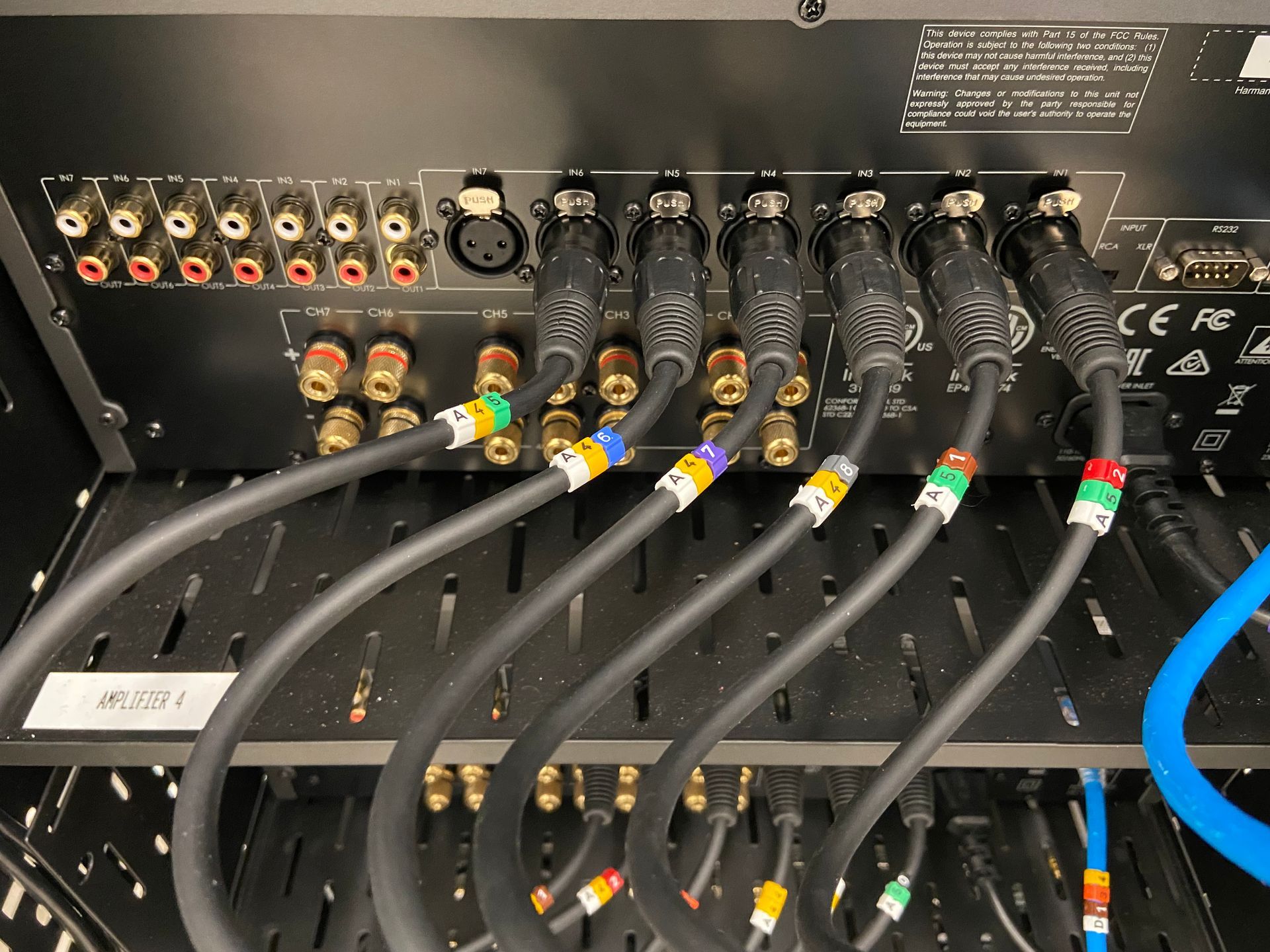 Rear view of an audio receiver with multiple connected cables, color-coded and labeled, inside a black rack.