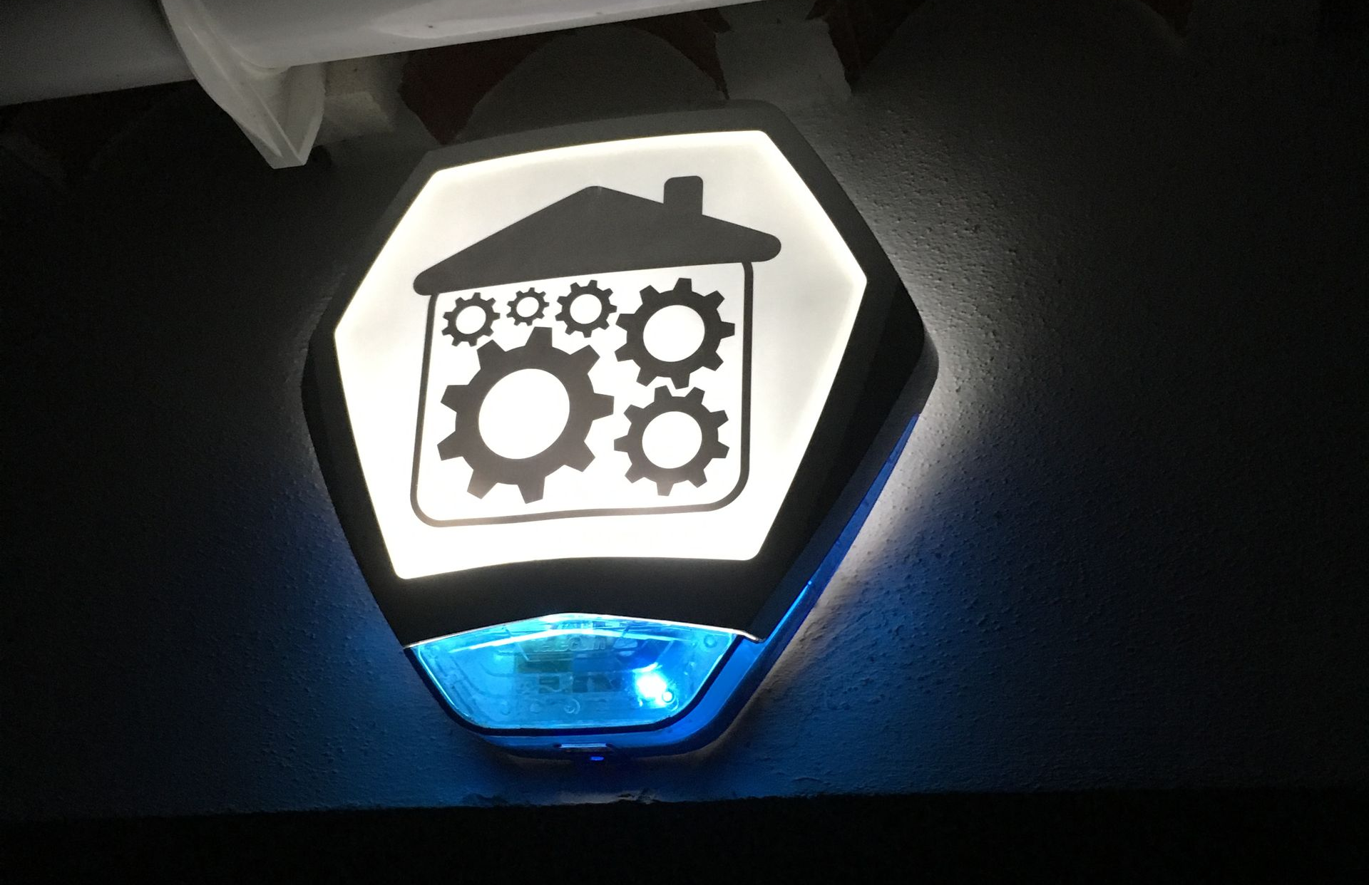 Intruder alarm sounder bell: white hexagon with a house and gears icon, blue light below.